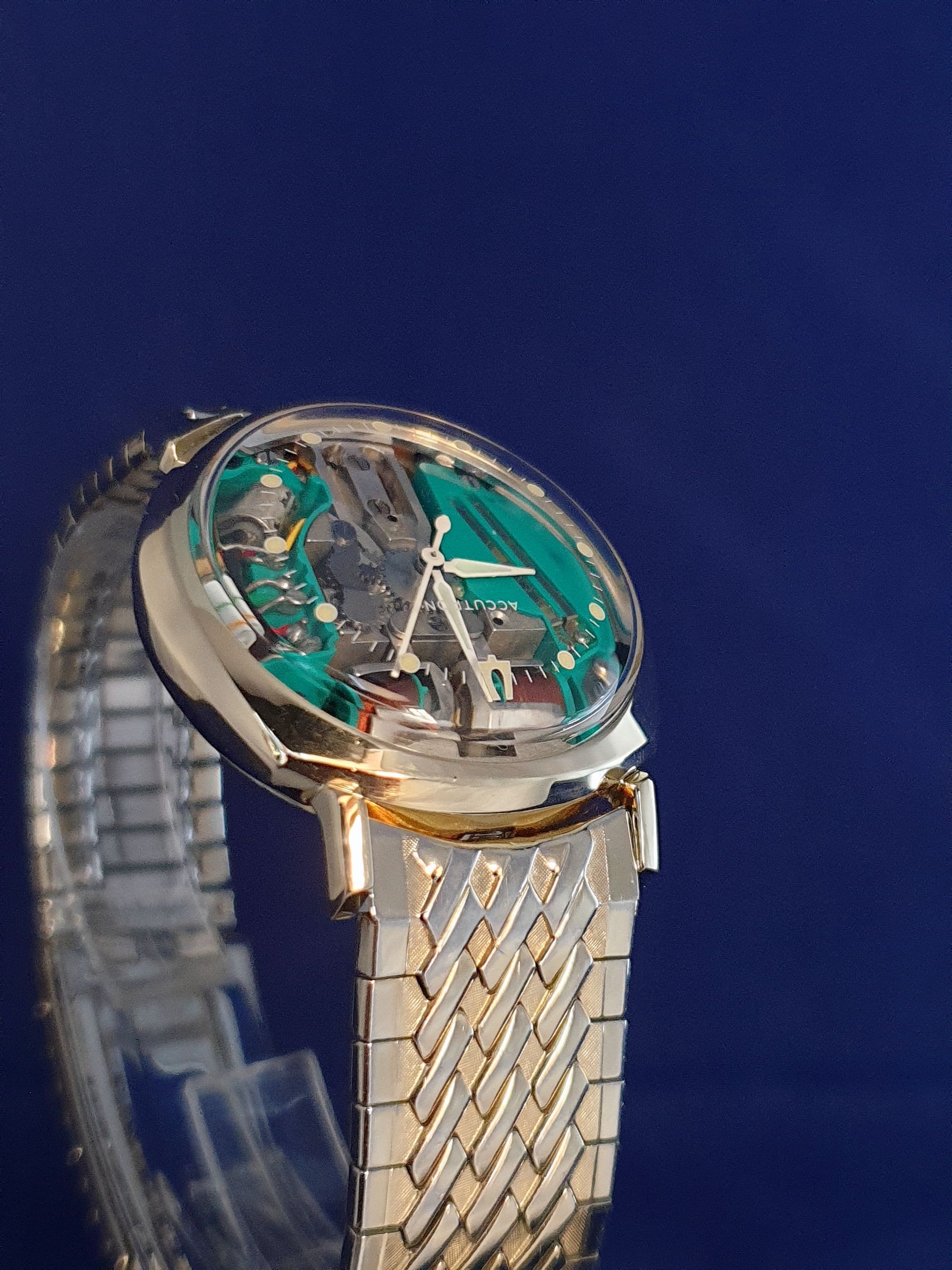 Luxury wristwatch with teal dial and gold accents on a silver woven bracelet, against a blue background