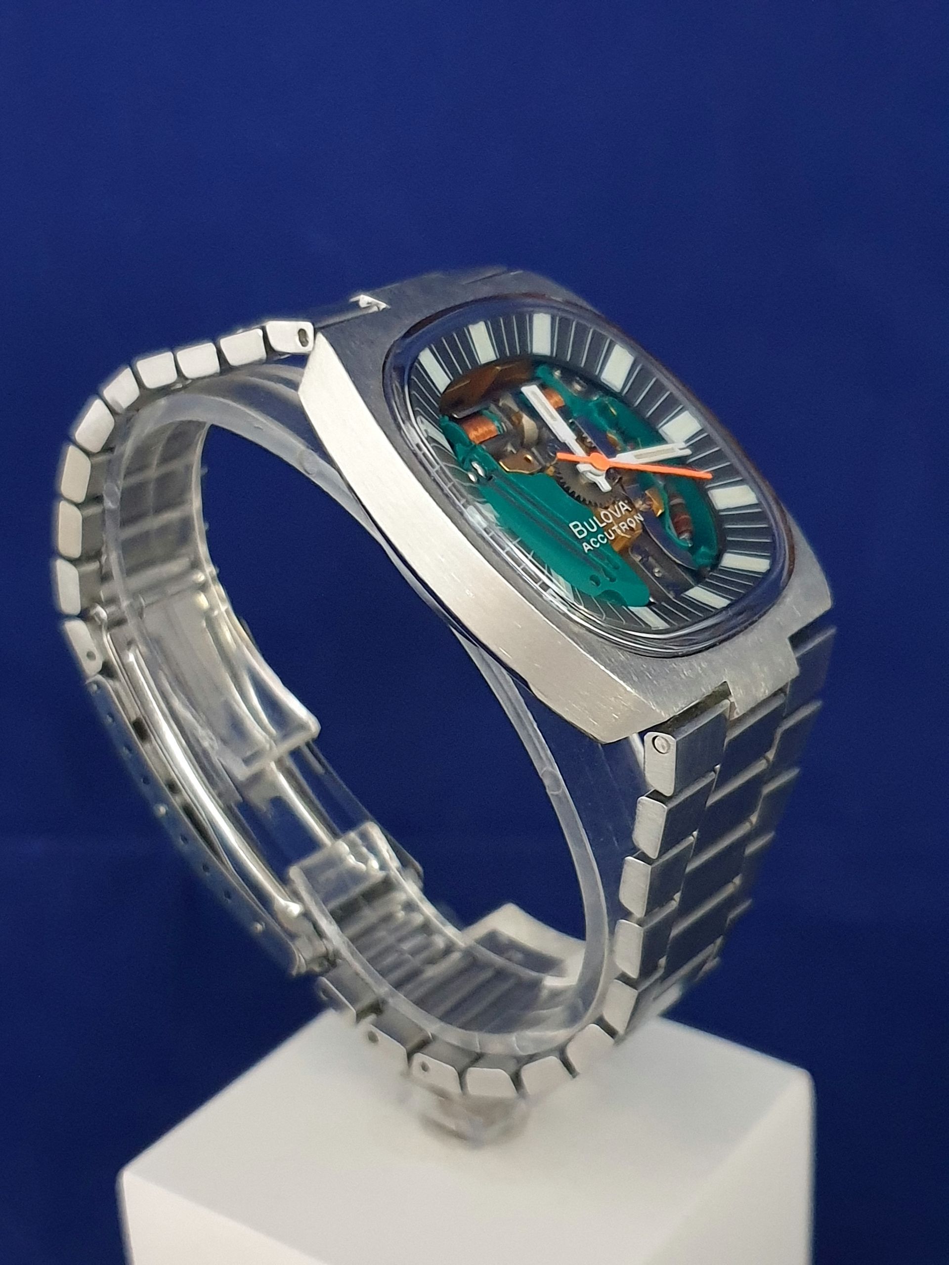 Transparent wristwatch with a teal face and silver band on a white display stand against a blue background