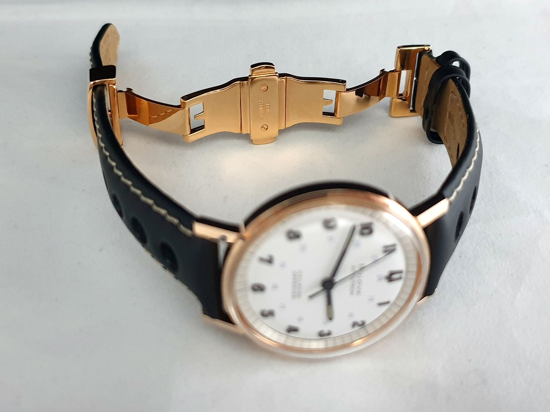 Black leather wristwatch with rose-gold case and white dial, open clasp shown on a light background
