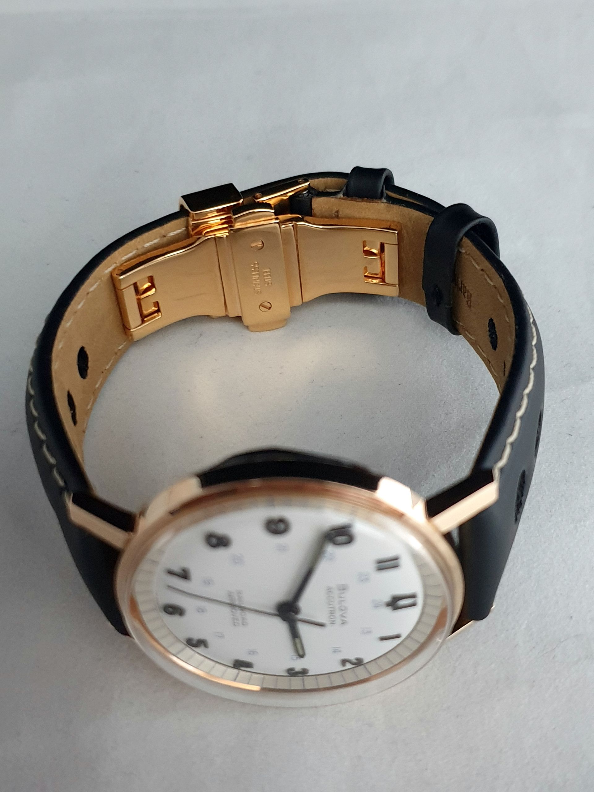Gold wristwatch with black strap, open clasp, showing a white dial on a light background