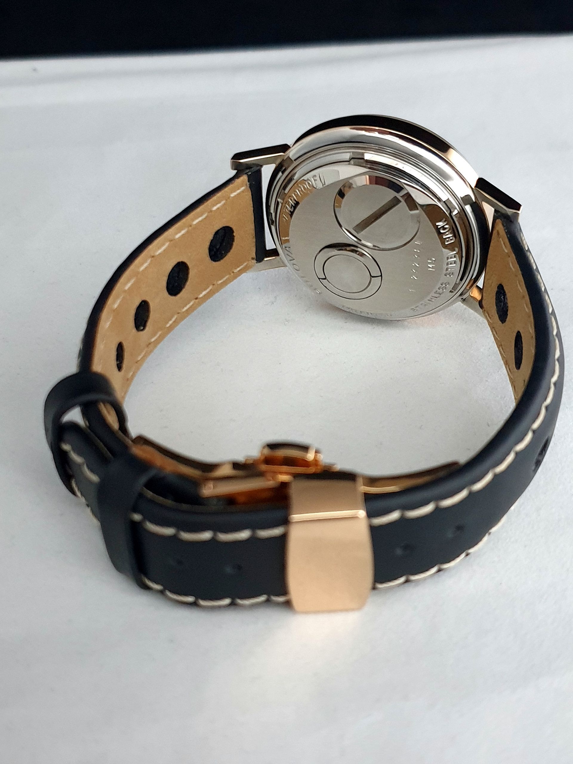 Black leather watch band with tan stitching and rose-gold clasp, showing the silver case back.