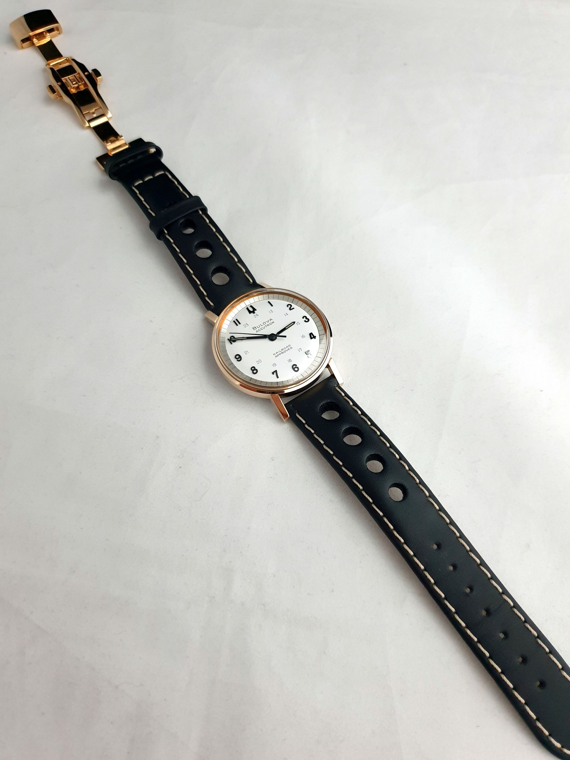 Black leather watch strap with gold buckle and a white-faced wristwatch on a light background