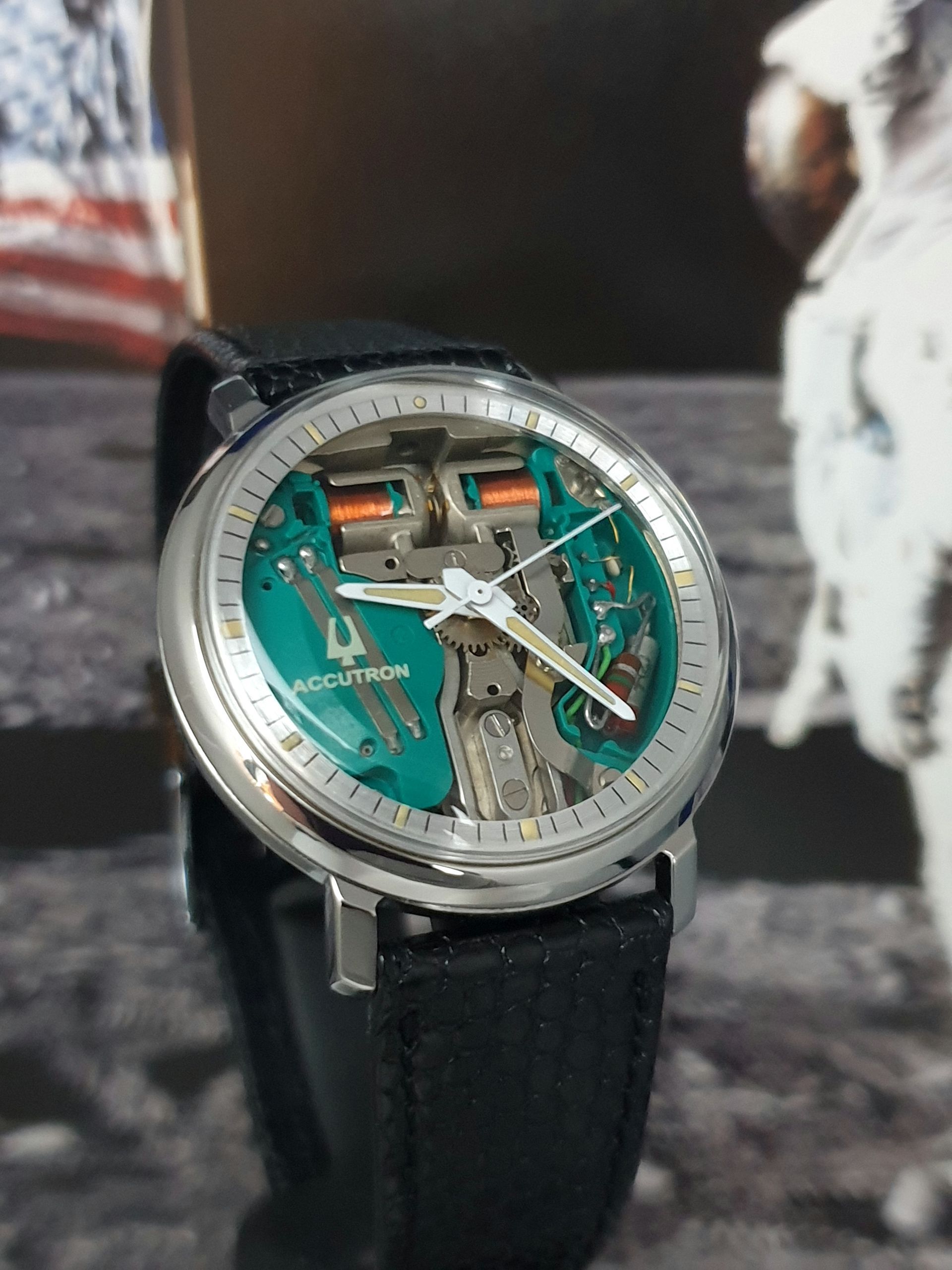 Close-up of a silver skeleton wristwatch with a green dial and black strap against a lunar astronaut backdrop