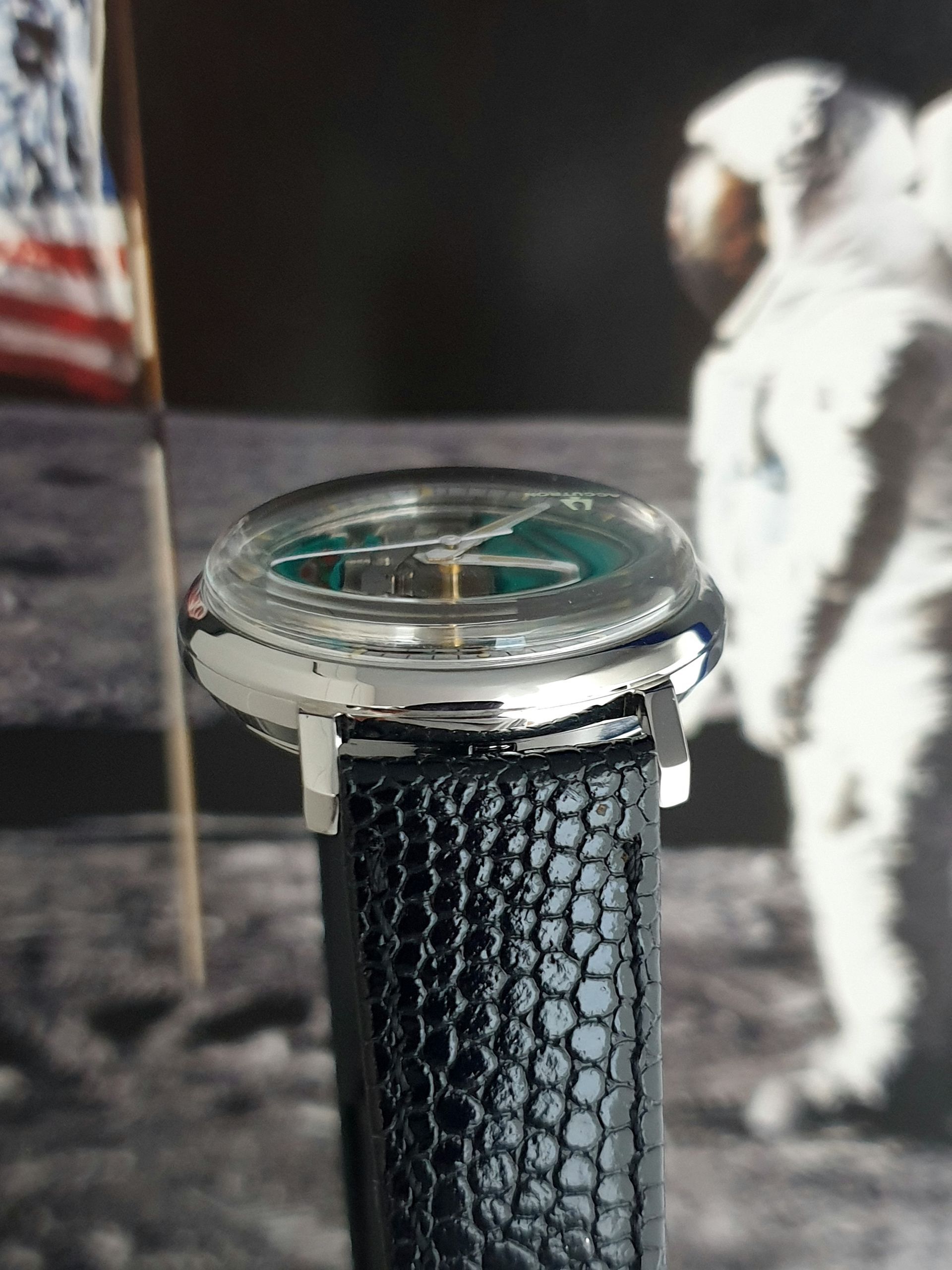 Close-up of a wristwatch with a black textured strap on a table, with a cat in the background