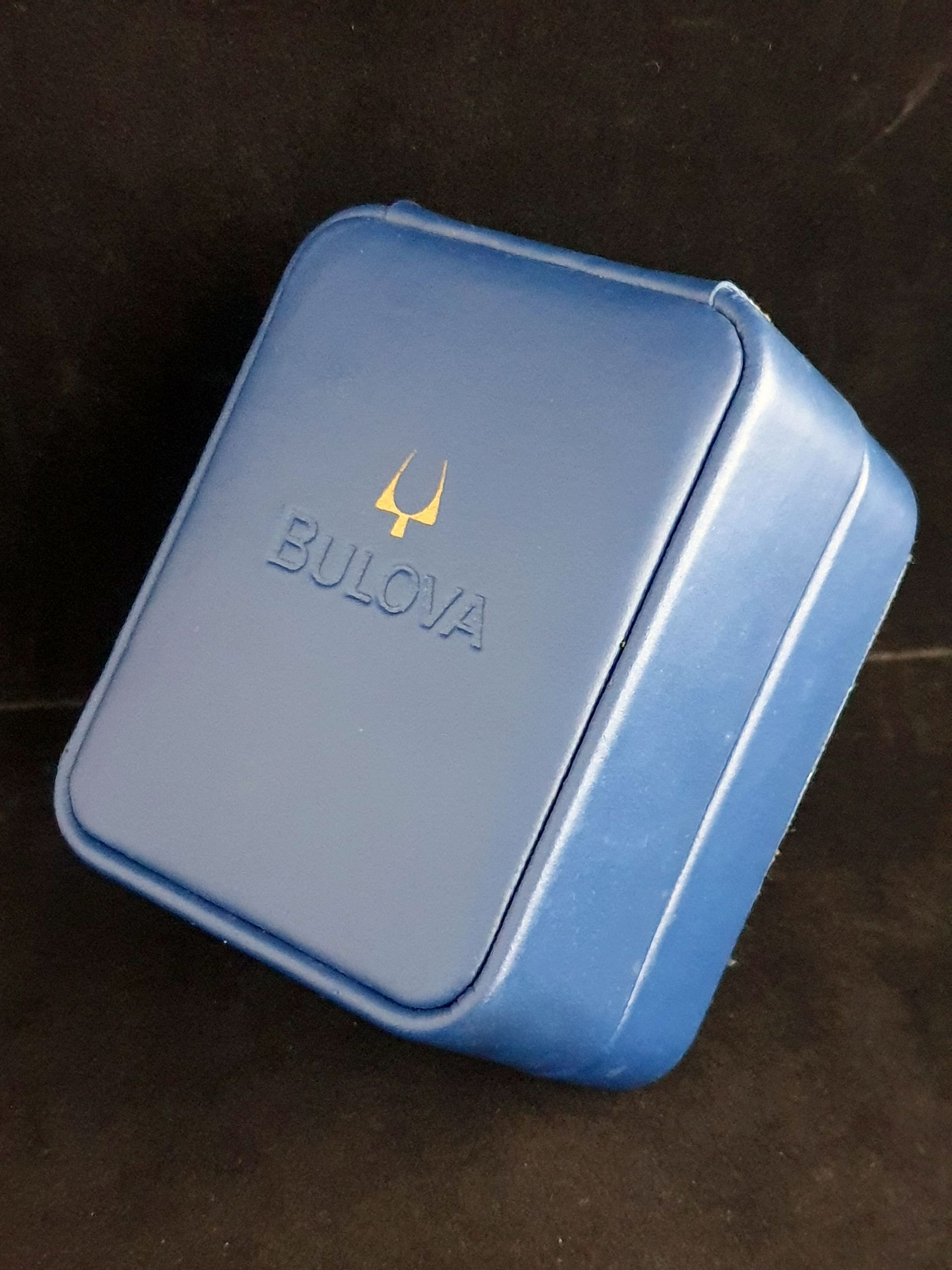 Blue square jewelry box with gold logo on a dark background