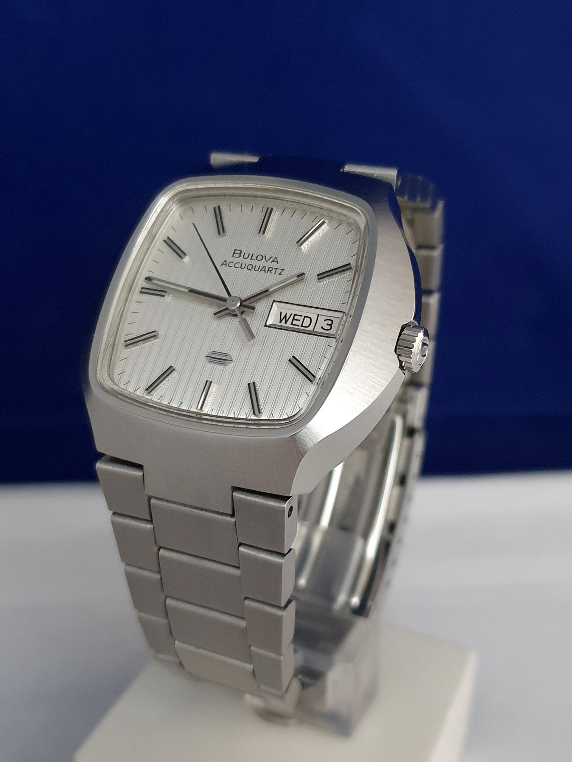 Silver metal wristwatch with a square face and day-date display on a blue background