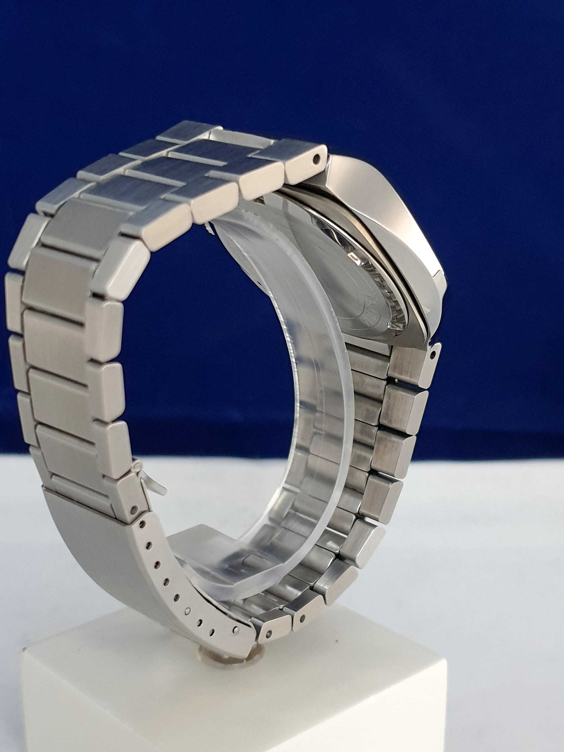 Silver metal wristwatch with open bracelet band on a white display stand against a blue background