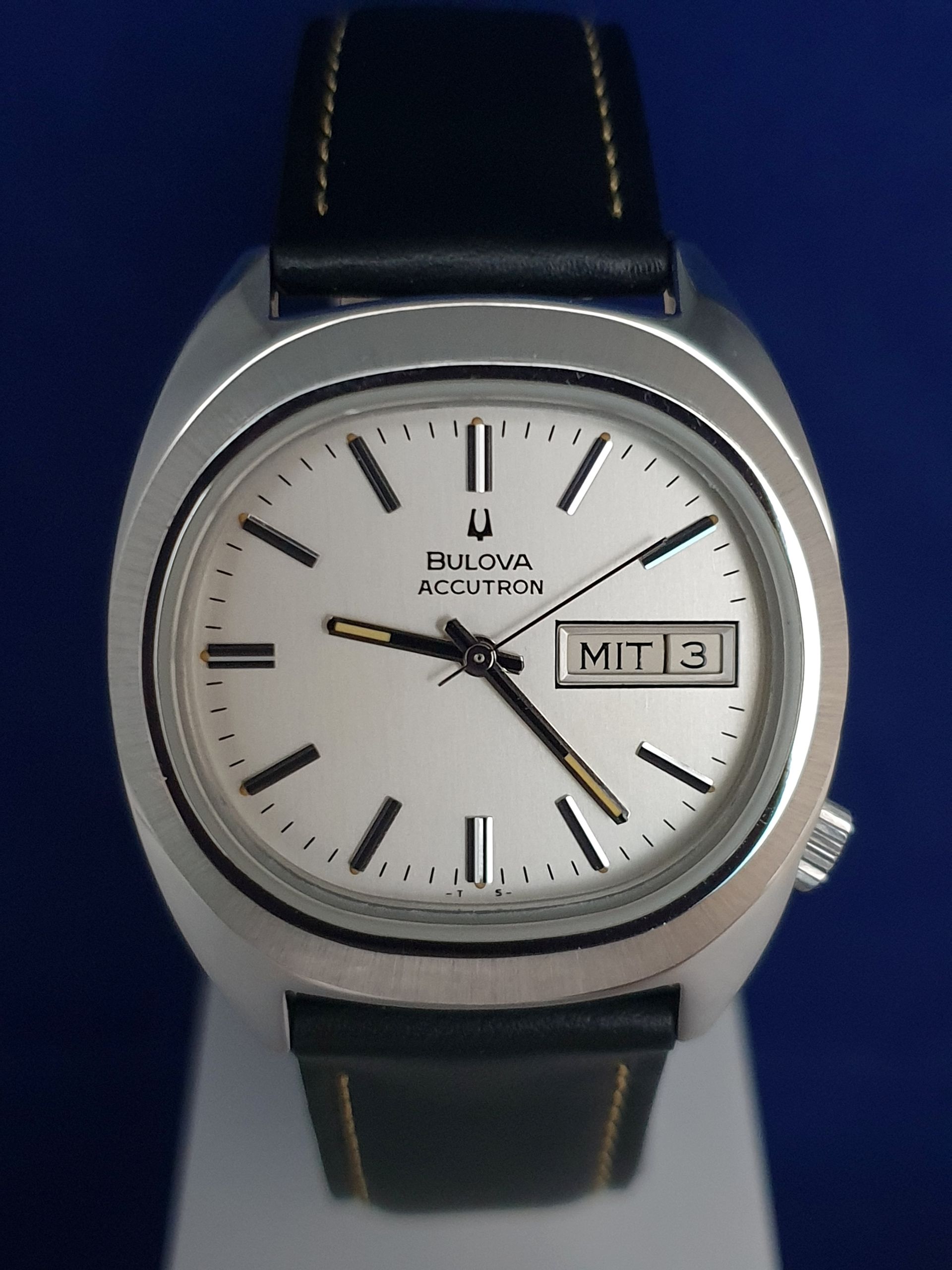 Bulova wristwatch with white face, black leather strap, and day-date display showing M/T 3.