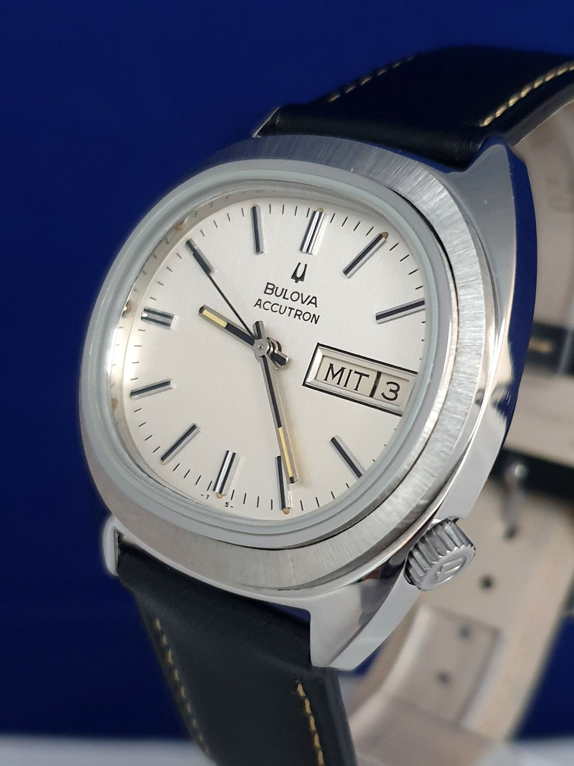 Silver wristwatch with white dial, black strap, and a date window on a blue background