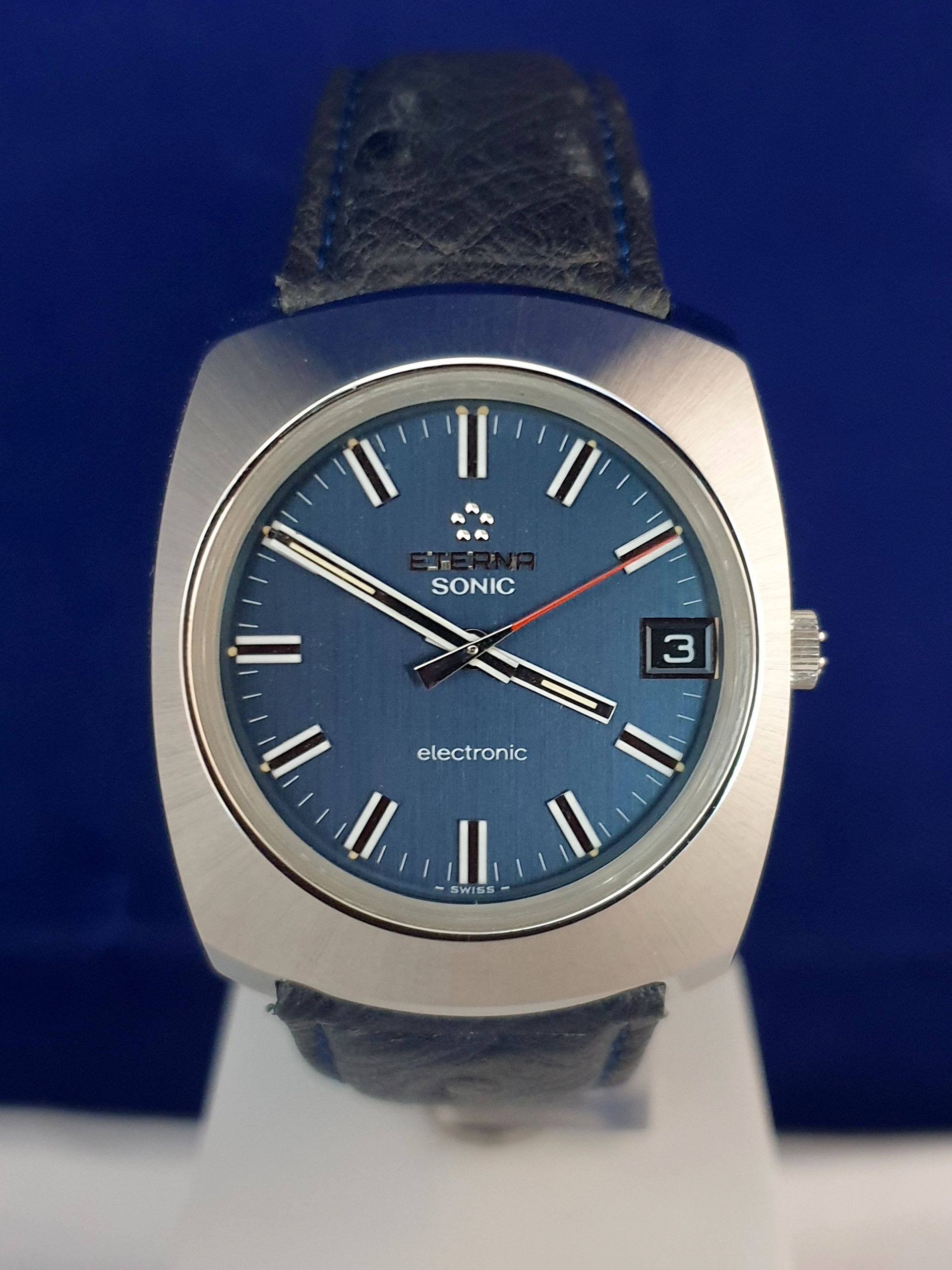 Blue-faced Sonic wristwatch with silver case, black strap, and date display on a stand