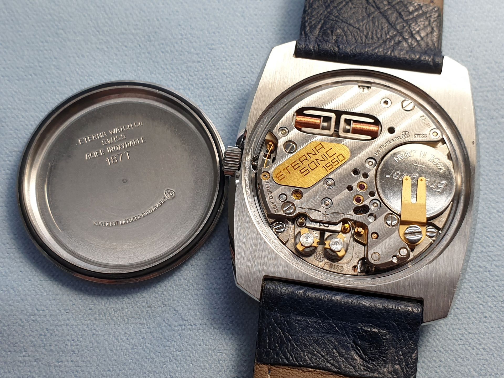 Open watch back showing movement and battery, with detached case back beside it on a blue surface