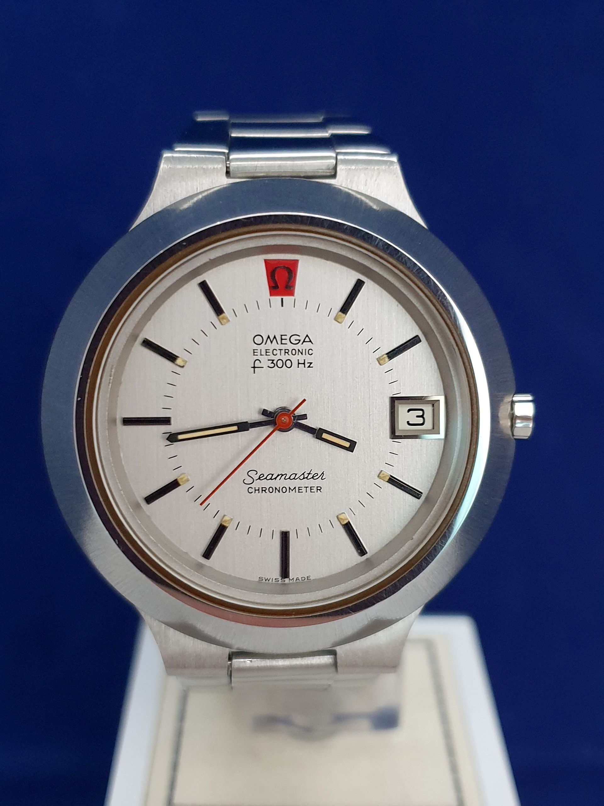Omega Seamaster watch with white dial, silver band, and orange seconds hand on a display stand