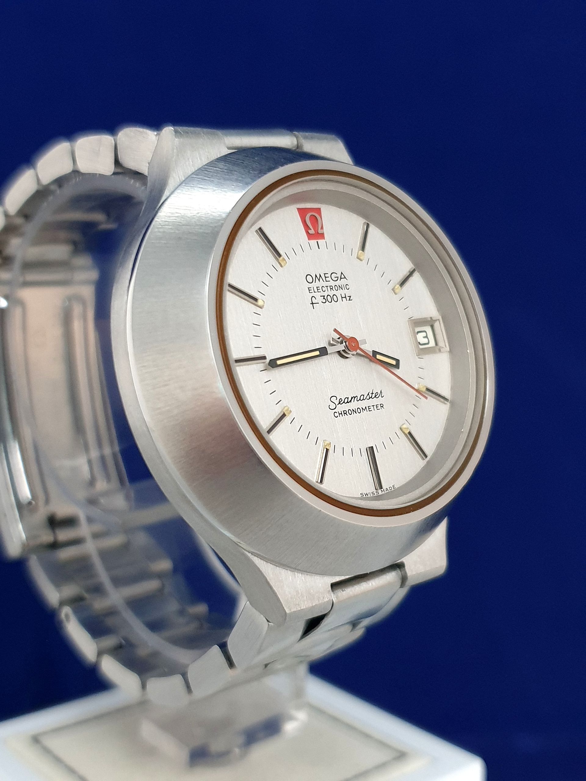 Silver Omega Constellation wristwatch with white dial on a clear stand against a blue background