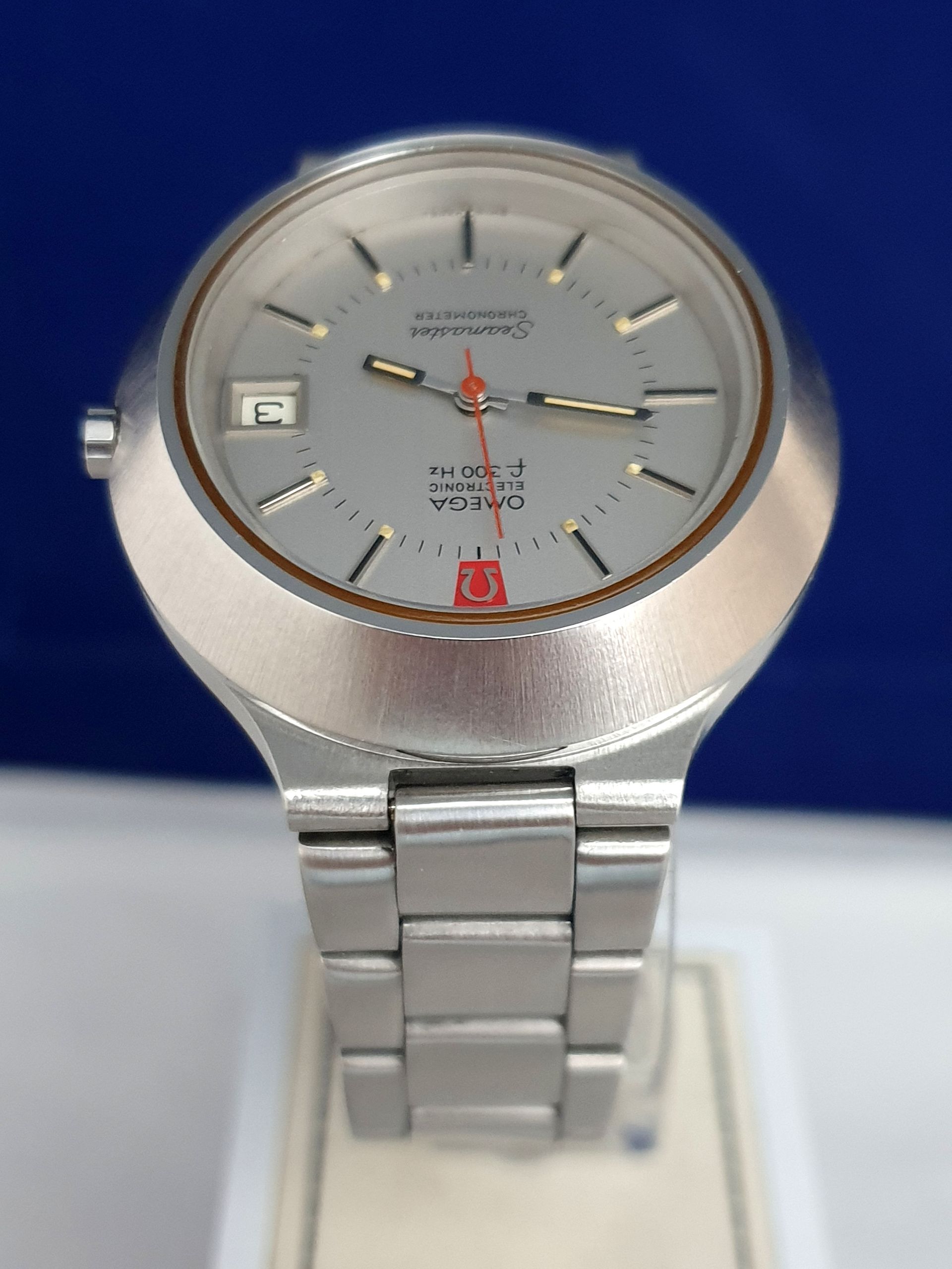 Silver wristwatch with gray dial and metal band on a stand, against a blue background