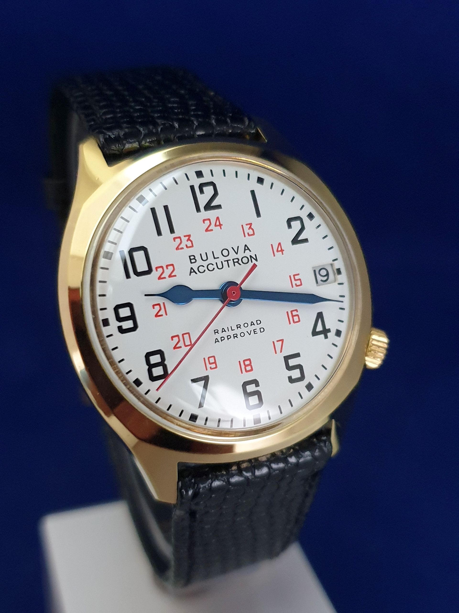 Bulova wristwatch with white dial, gold bezel, and black leather band on a blue background