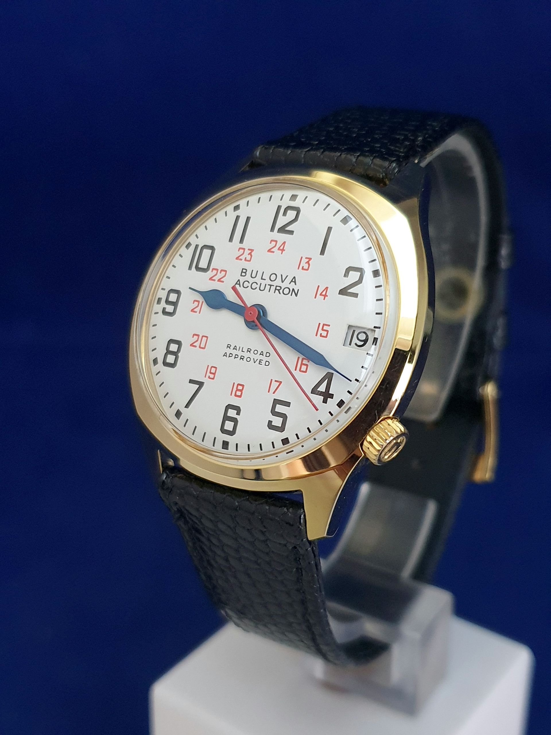 Gold-tone wristwatch with white dial and black strap on a display stand
