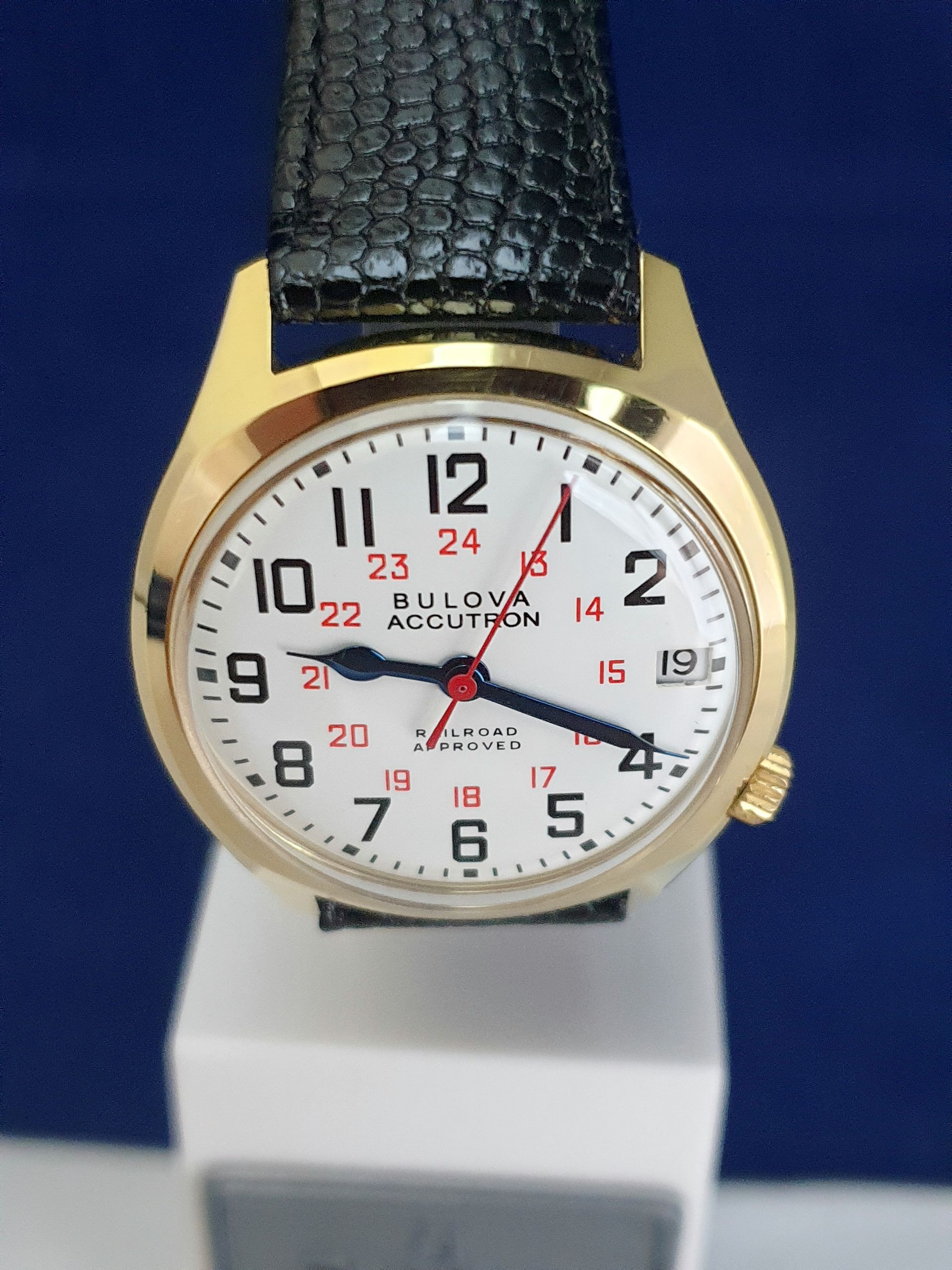 Gold wristwatch with white dial, black leather strap, and red 24-hour markings on a display stand