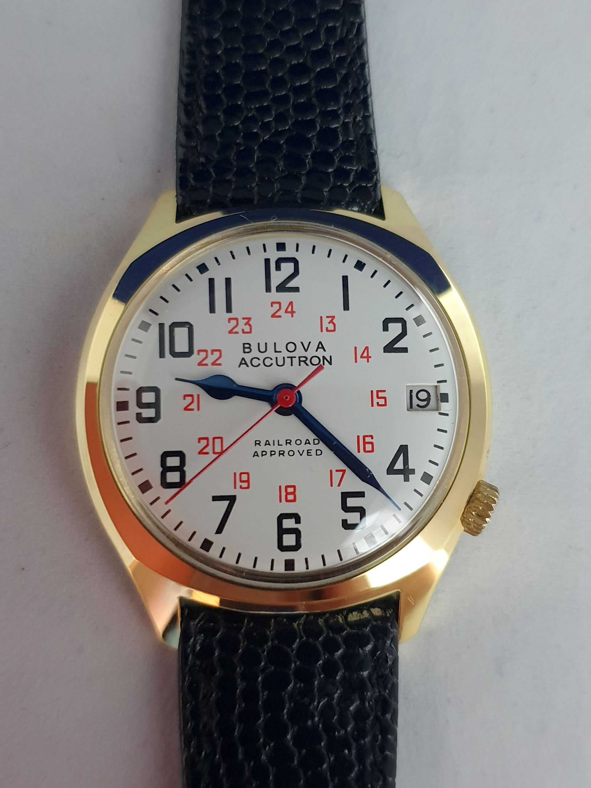 Gold wristwatch with black textured band and white face showing Arabic numerals and red 24-hour markings