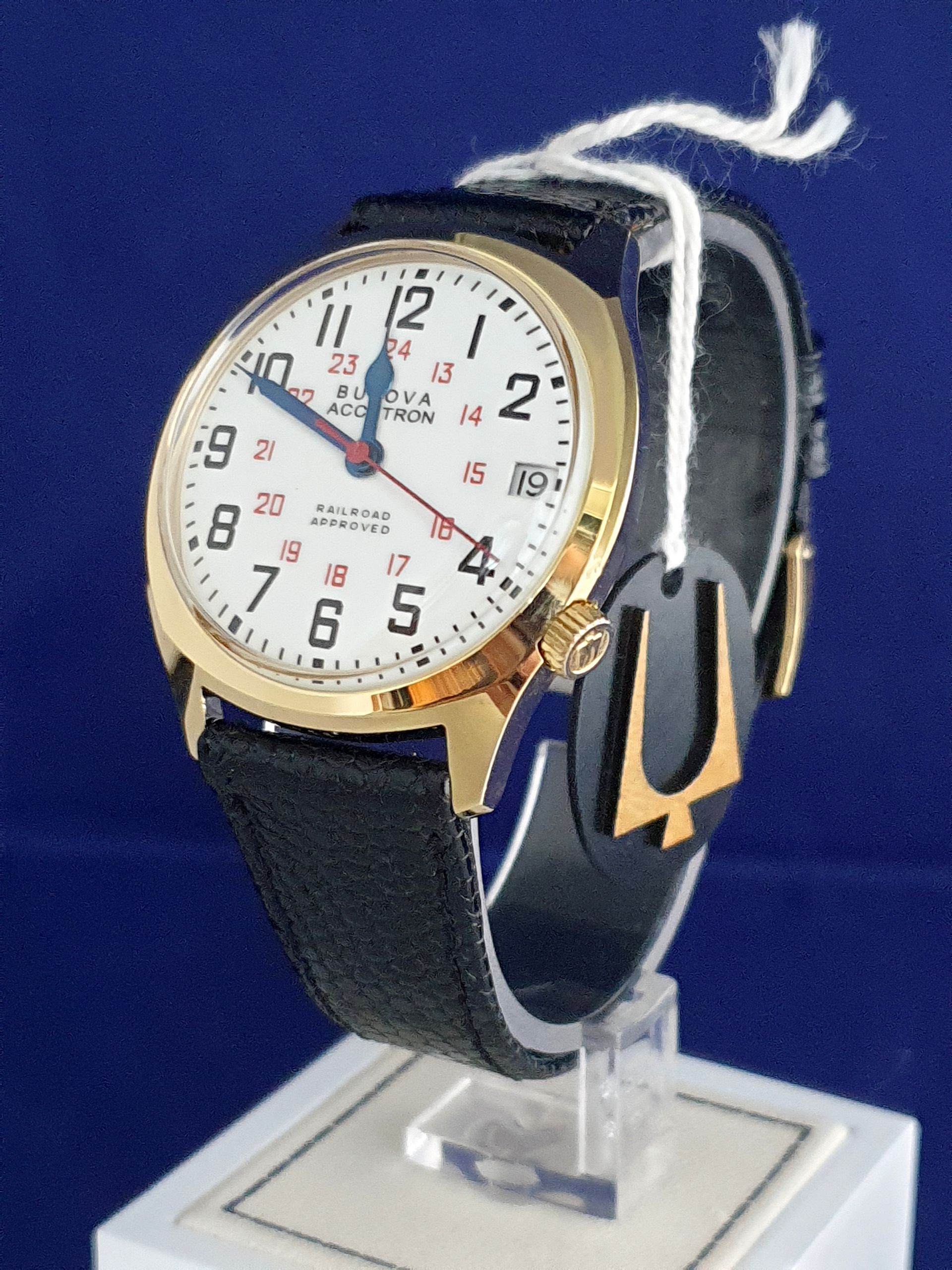 Luxury wristwatch with white dial, gold bezel, and black strap on a clear display stand
