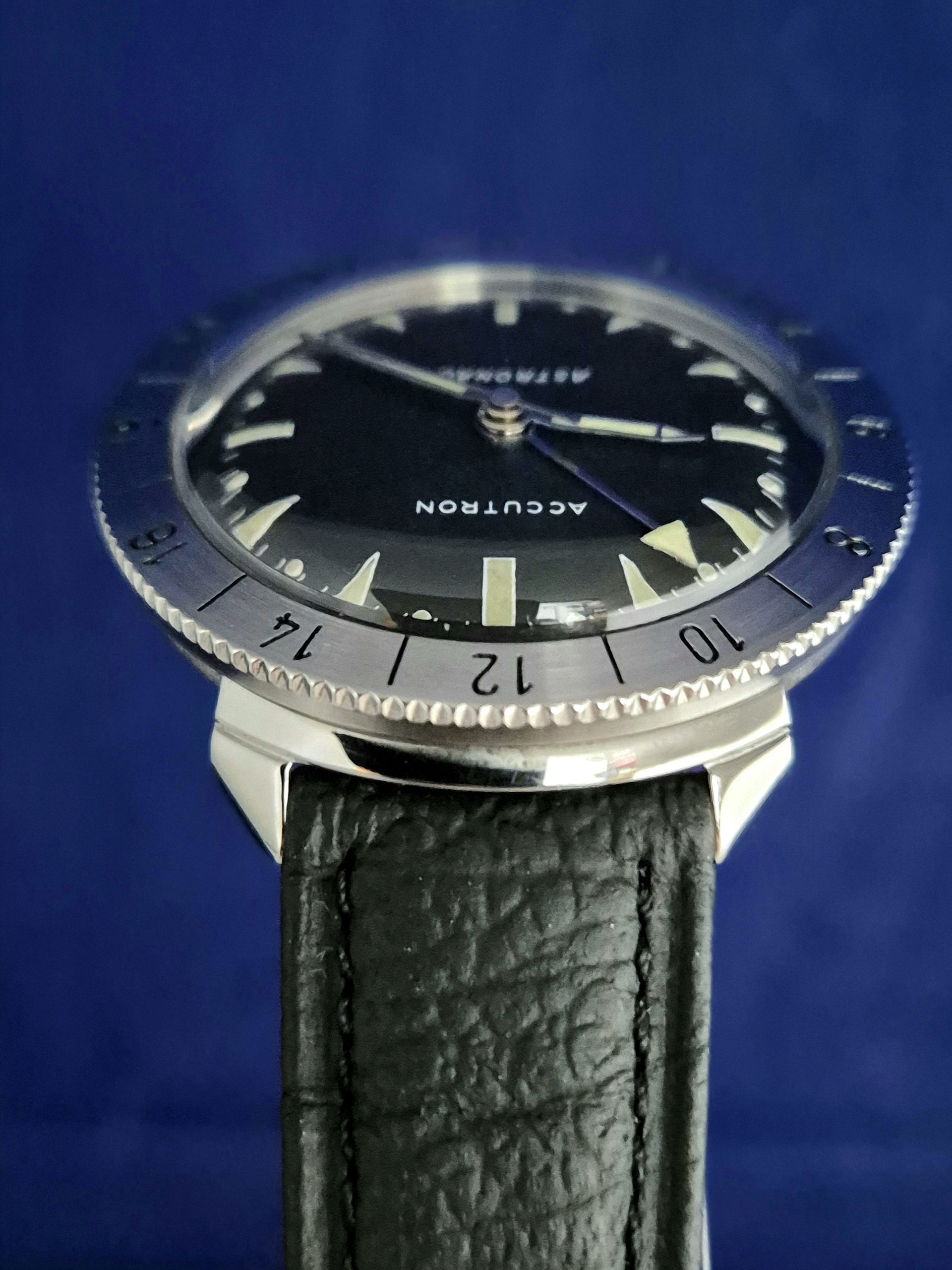 Close-up of a silver dive watch with black dial and black leather strap on a blue background