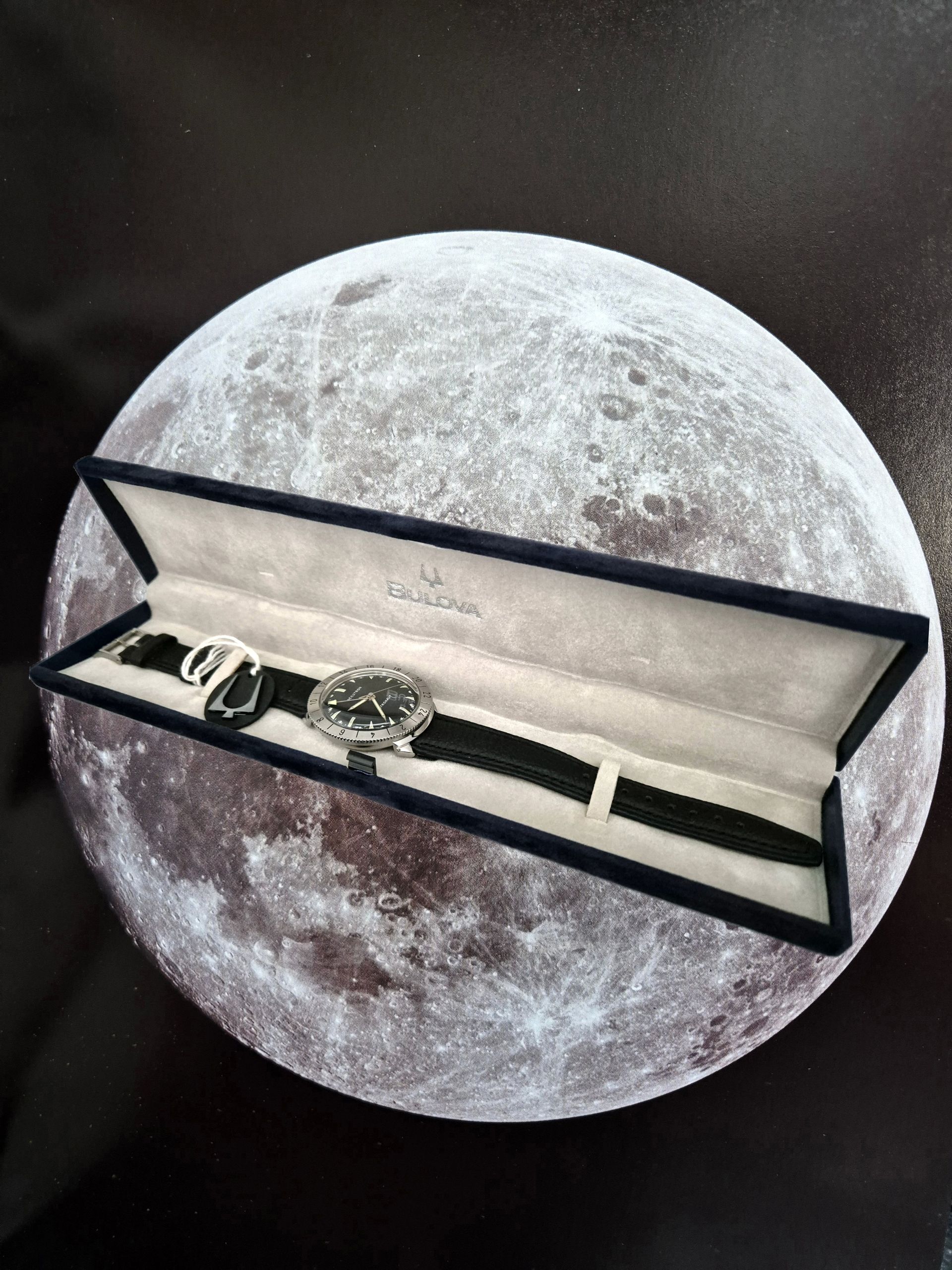 Open watch display case on a moon-themed background, showing a wristwatch inside.