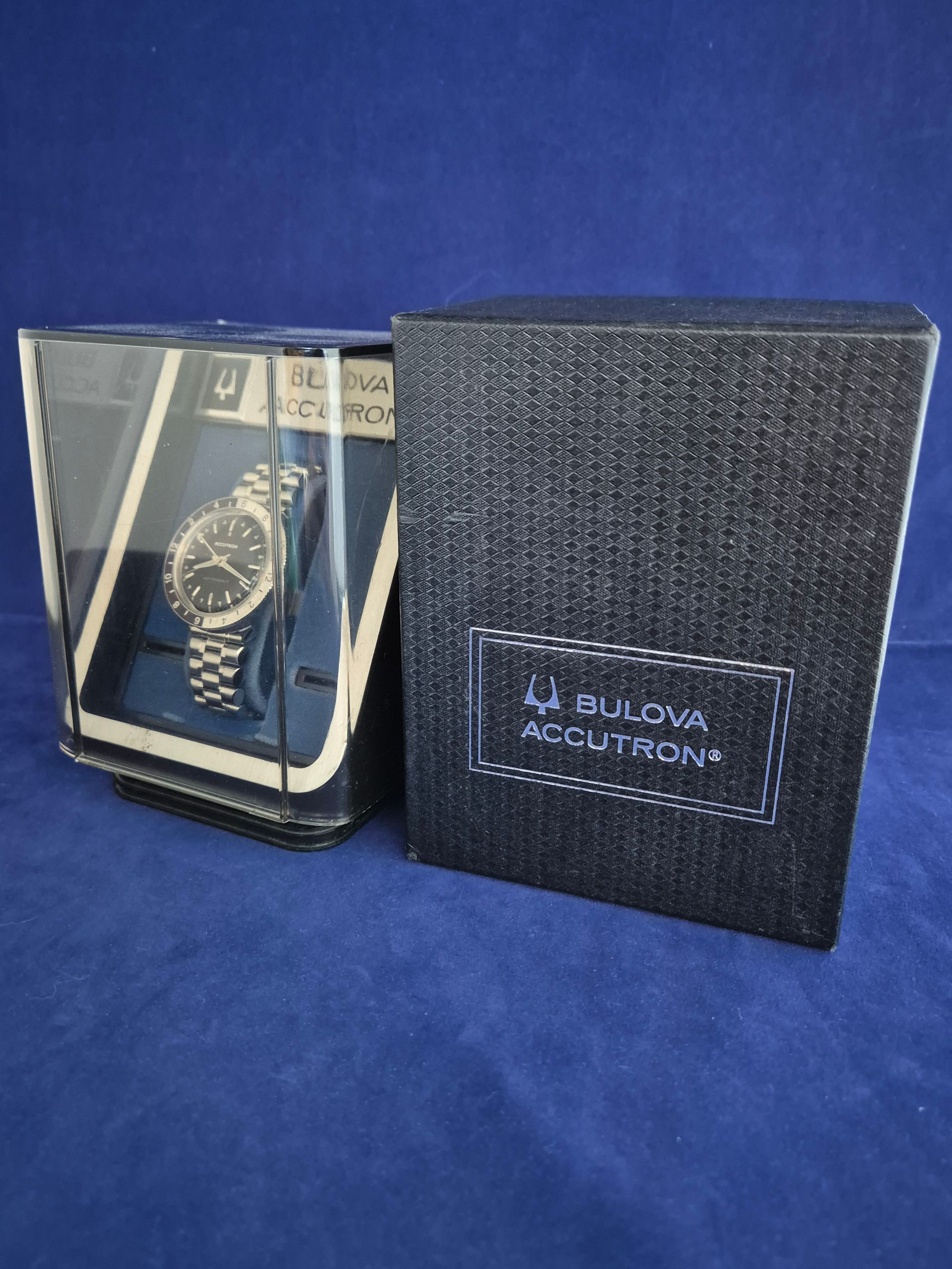 Bulova Accutron watch in display case beside black Bulova Accutron box on blue background