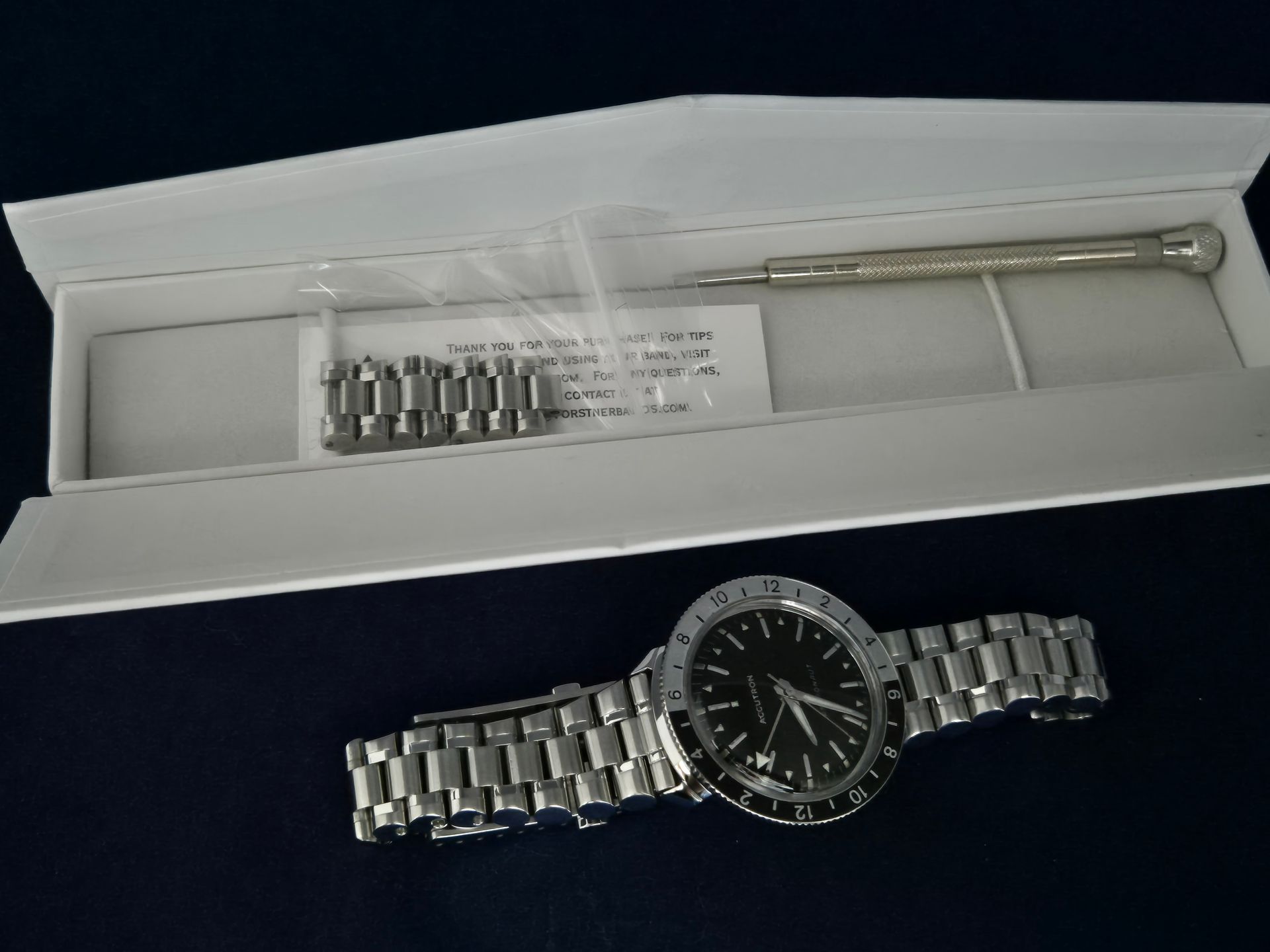 Silver wristwatch and extra links displayed in an open white box on a dark background