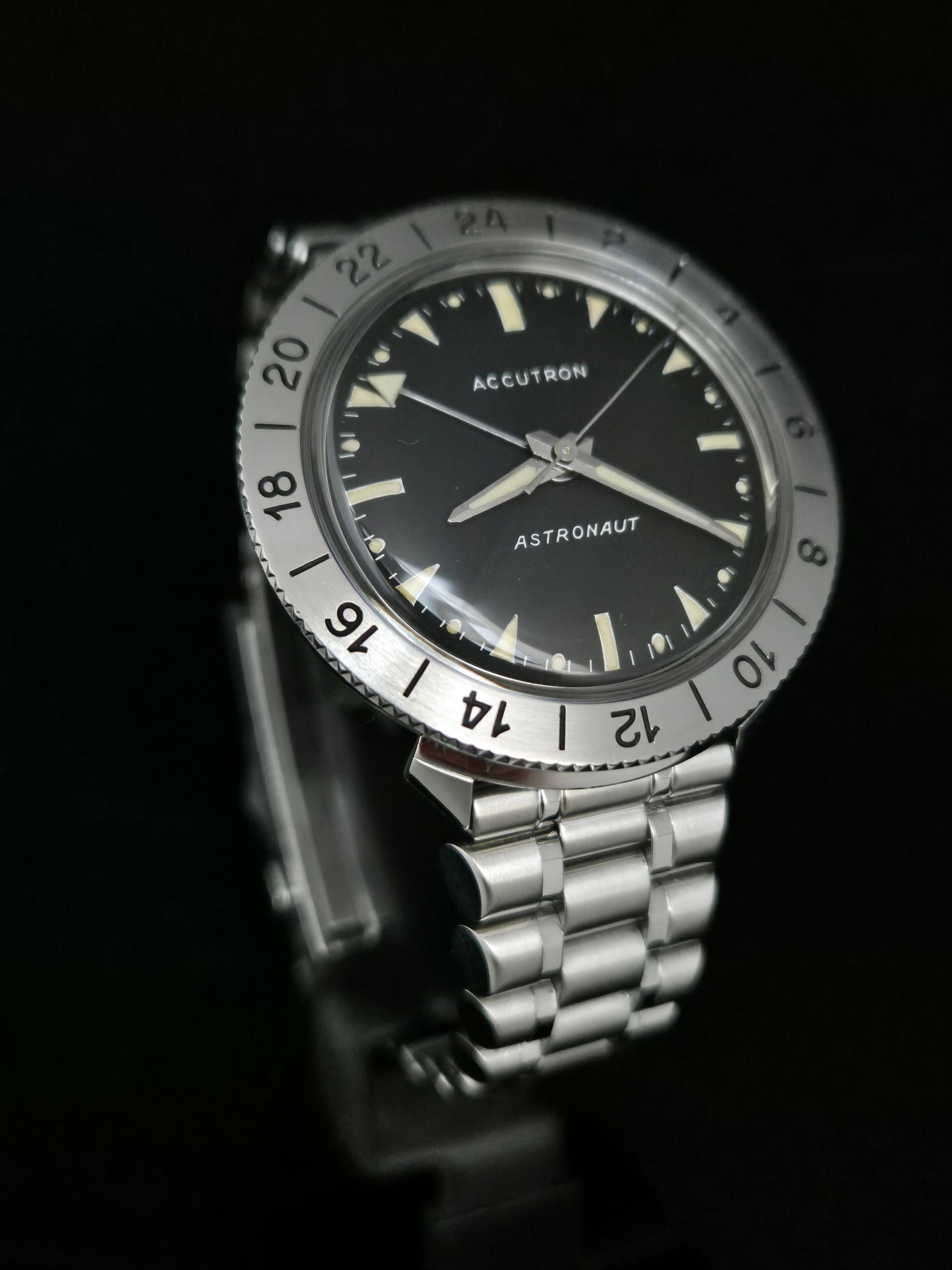 Silver wristwatch with black dial and white bezel, shown against a dark background.