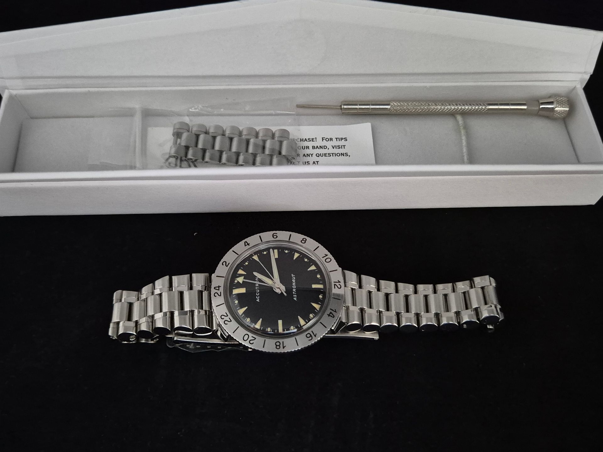 Silver wristwatch with metal band in an open display box, with extra links and a tool above it