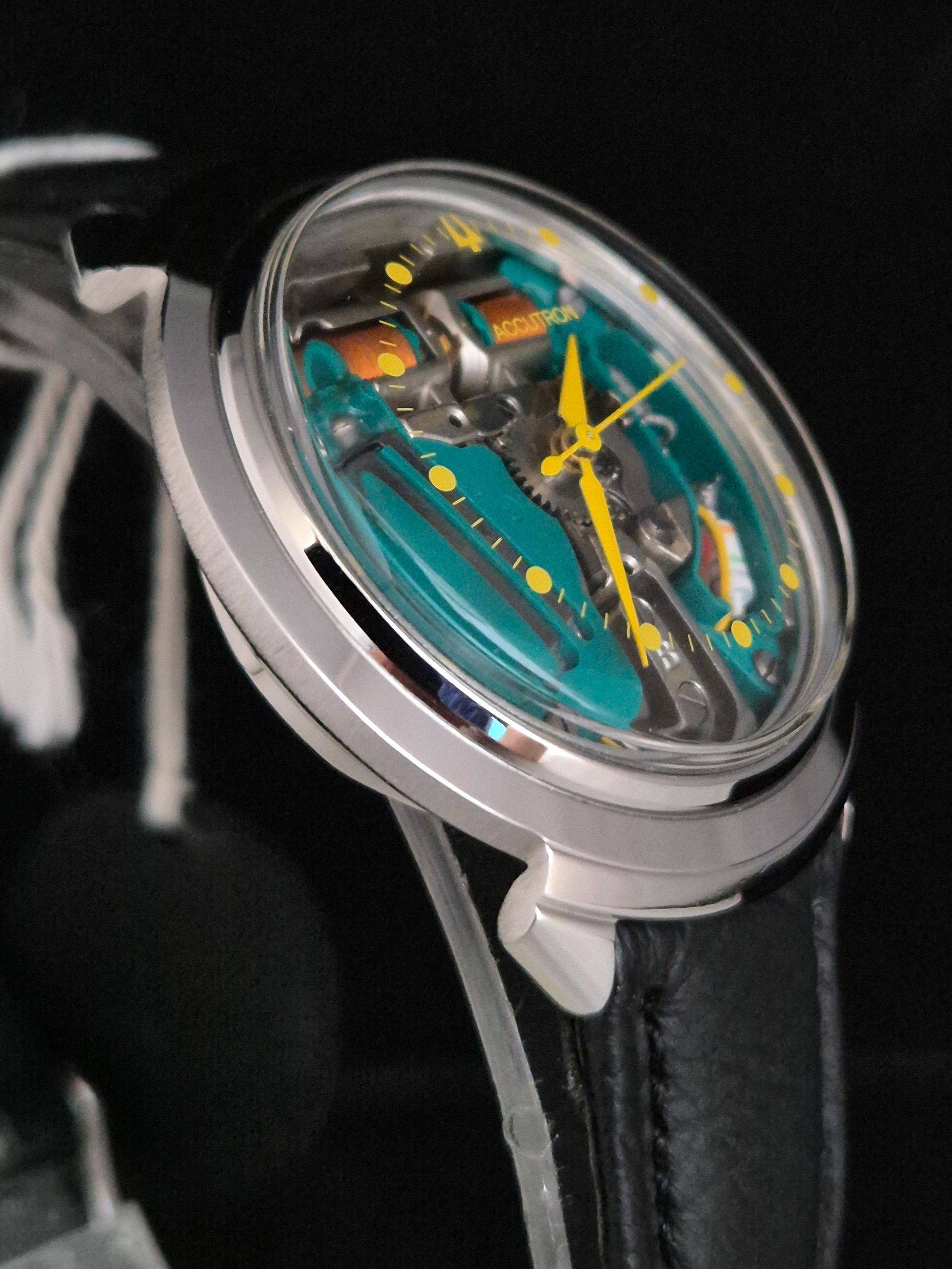 Close-up of a transparent mechanical wristwatch with teal and yellow details on a black strap