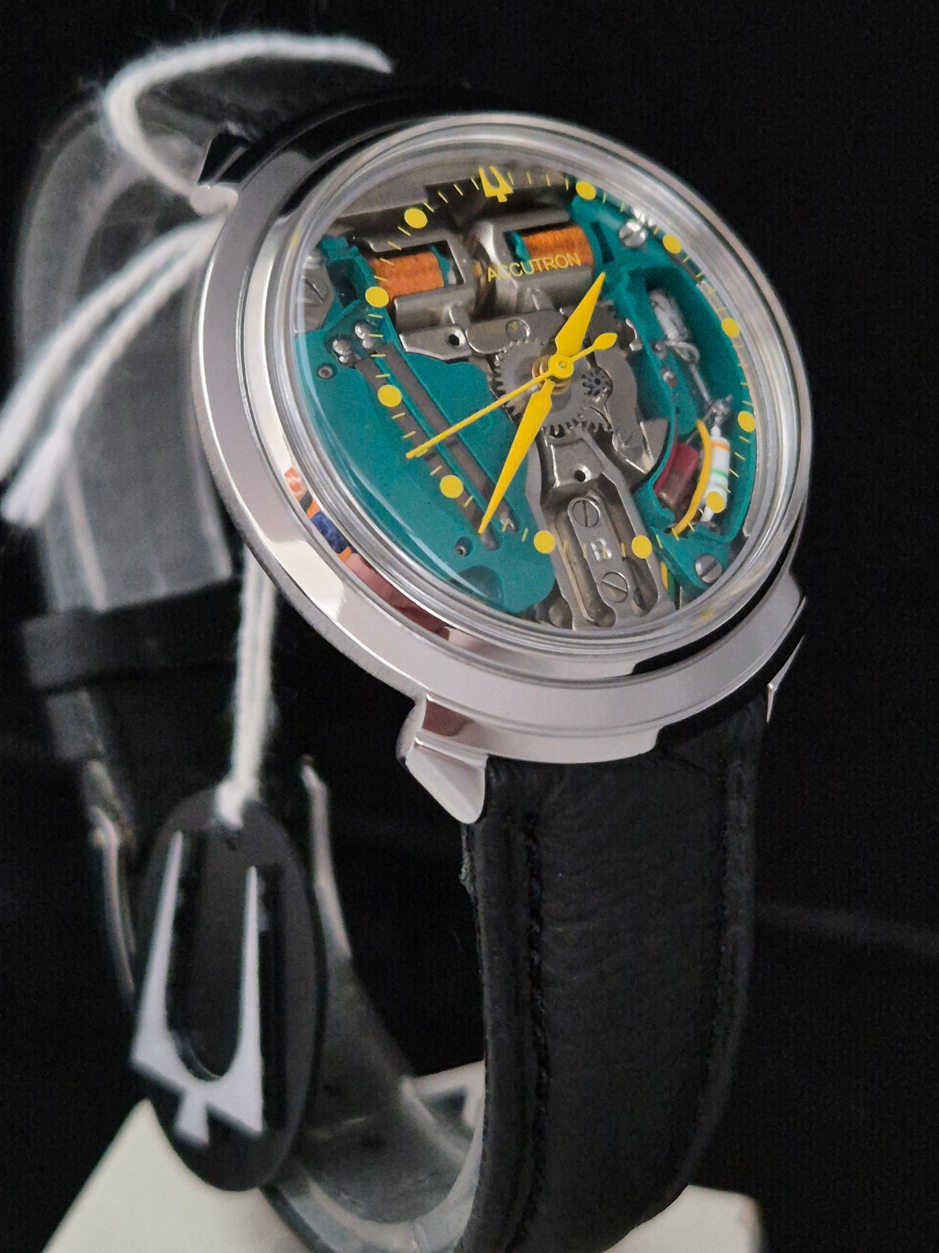 Skeleton wristwatch with teal dial, yellow hands, and black leather strap on a gray background