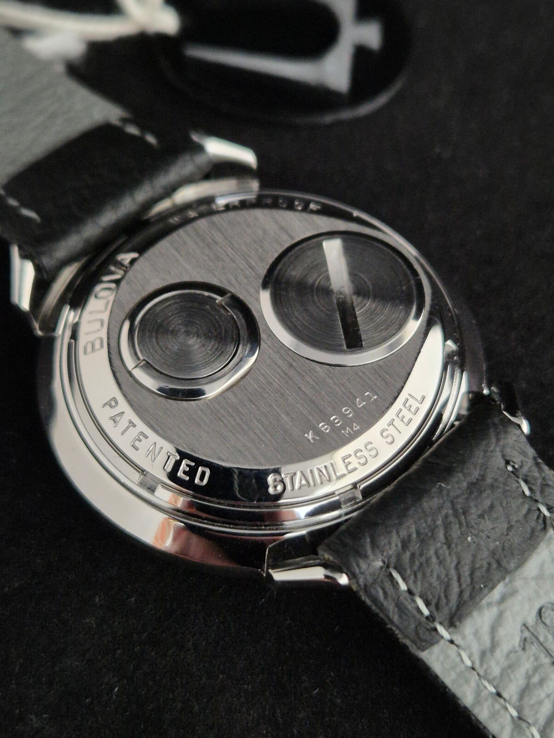 Close-up of a silver watch back with black leather strap, showing two circular engraved details.