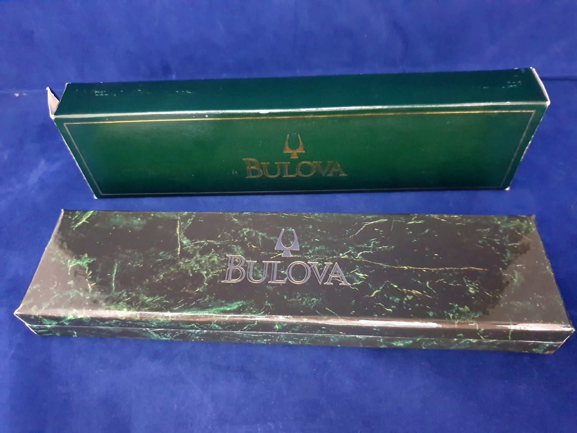 Two Bulova watch boxes, one green and one marbled purple, on a blue cloth background