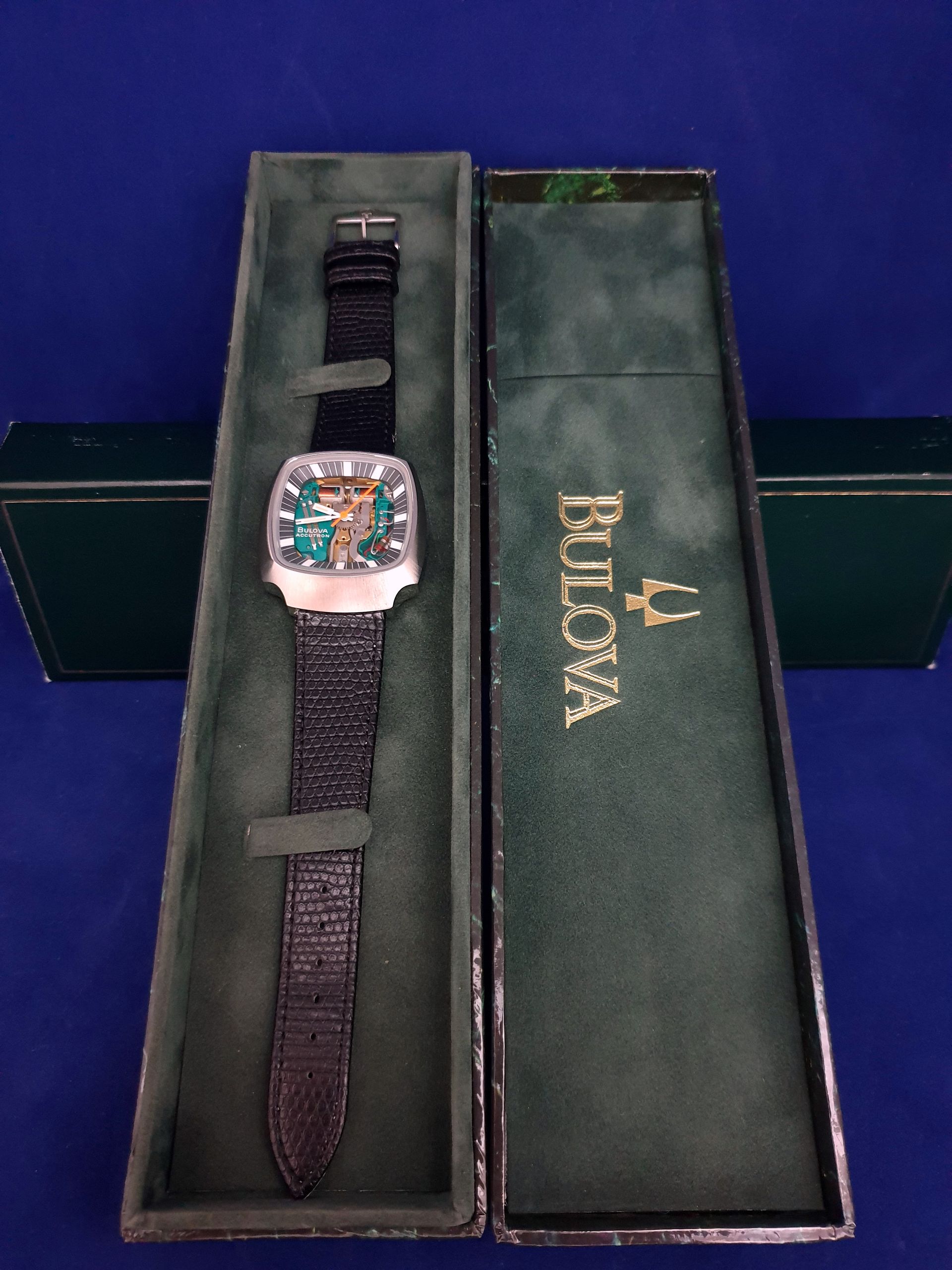Bulova wristwatch in open box, with dark strap and rose gold bezel on green case insert