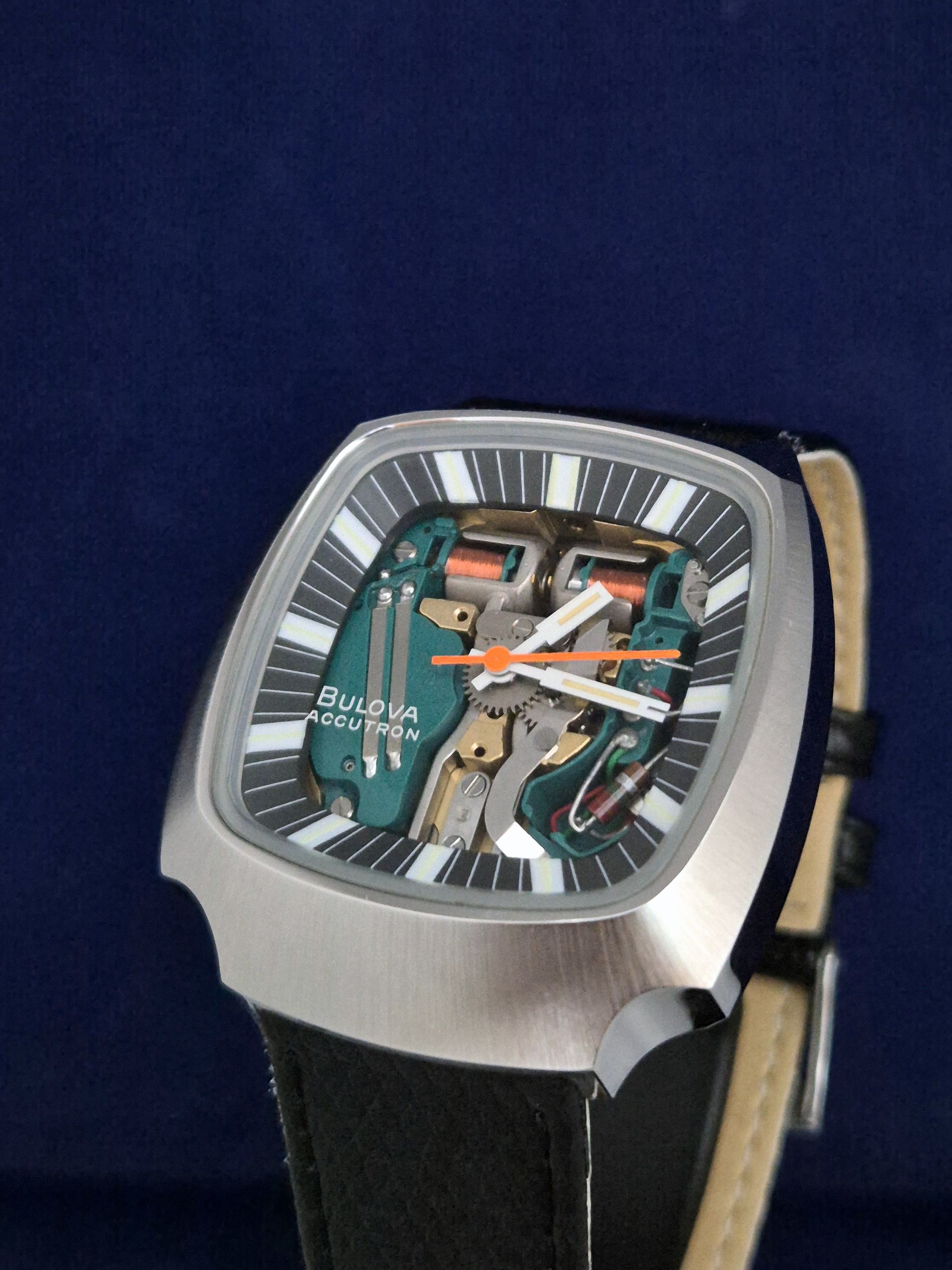 Silver wristwatch with a rectangular dial, orange and green hands, and a black strap on a dark blue background