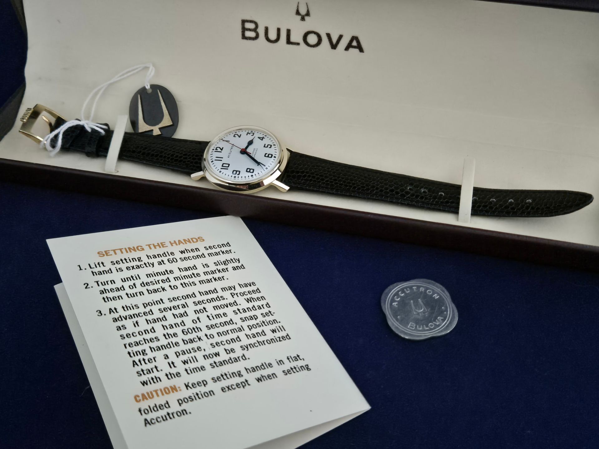 Bulova watch with black strap on a white display box, with warranty card and coin nearby