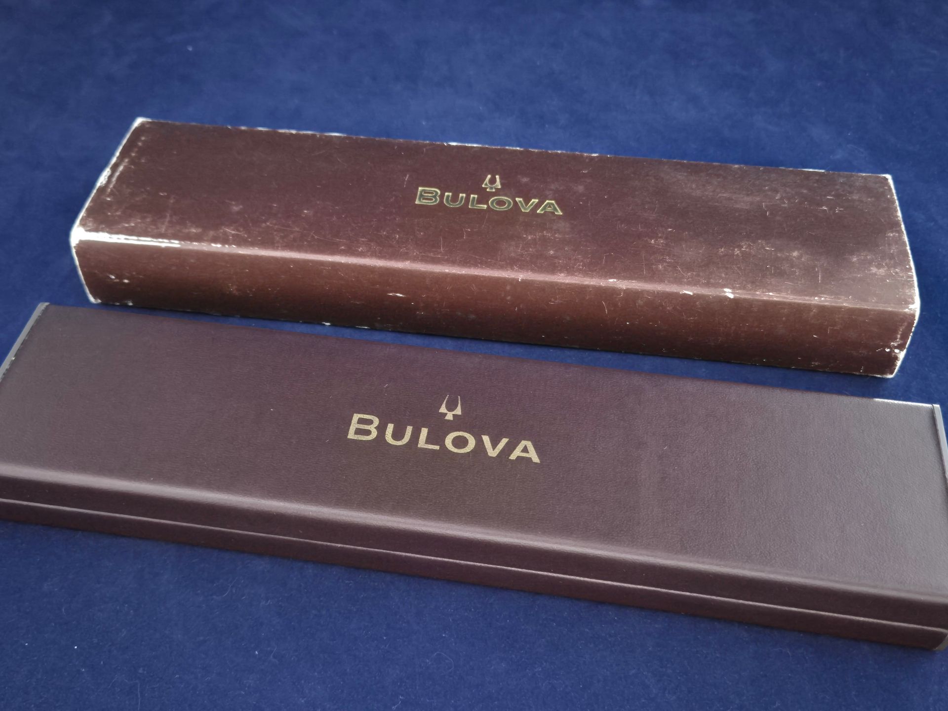 Two Bulova watch boxes, one brown and one dark blue, stacked on a blue surface.