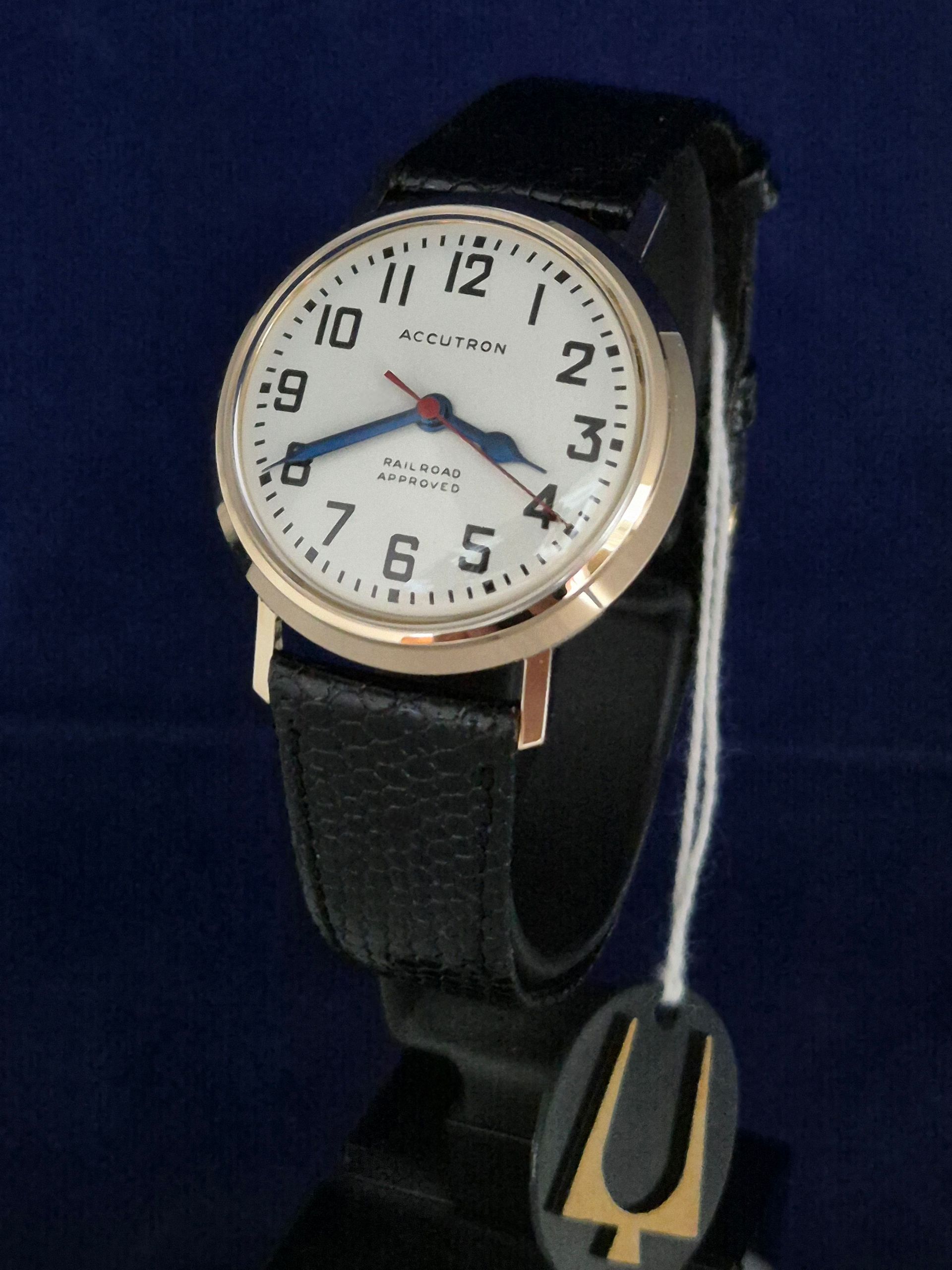 Black wristwatch with white dial and black strap, shown with a hanging tag on a blue background