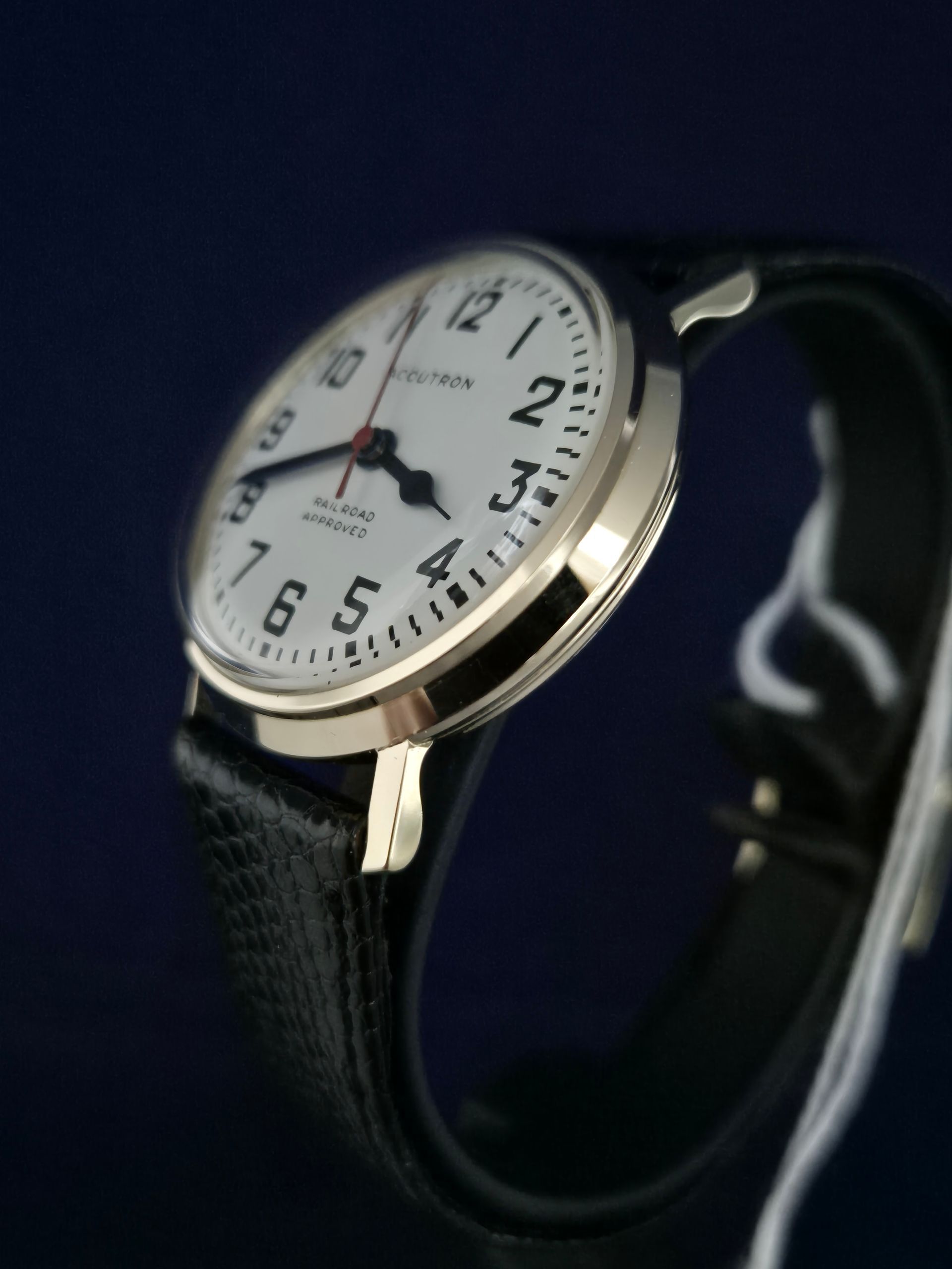 Analog wristwatch with white dial, gold bezel, and black strap on dark background