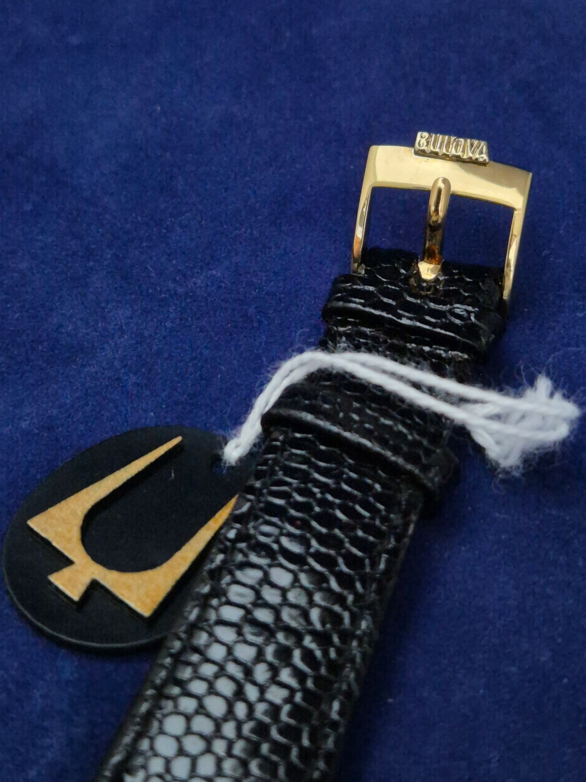 Black textured wristband with gold buckle and black-and-gold emblem on blue background