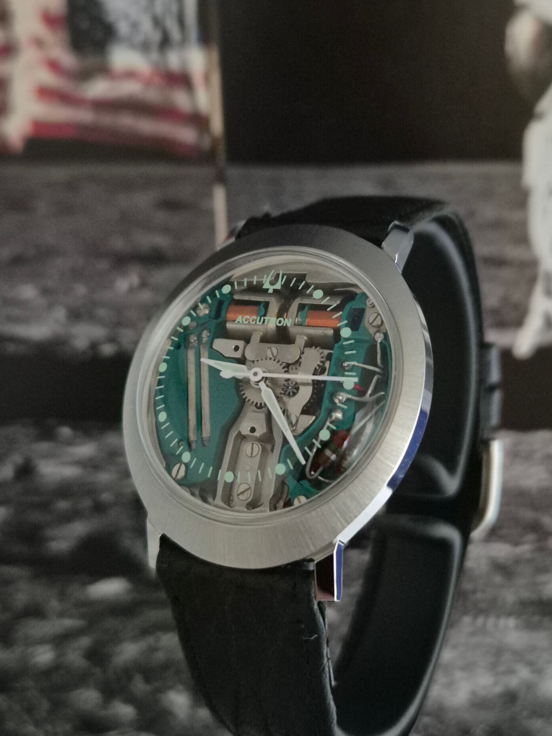 Transparent skeleton wristwatch with black strap on a dark surface, showing teal internal components