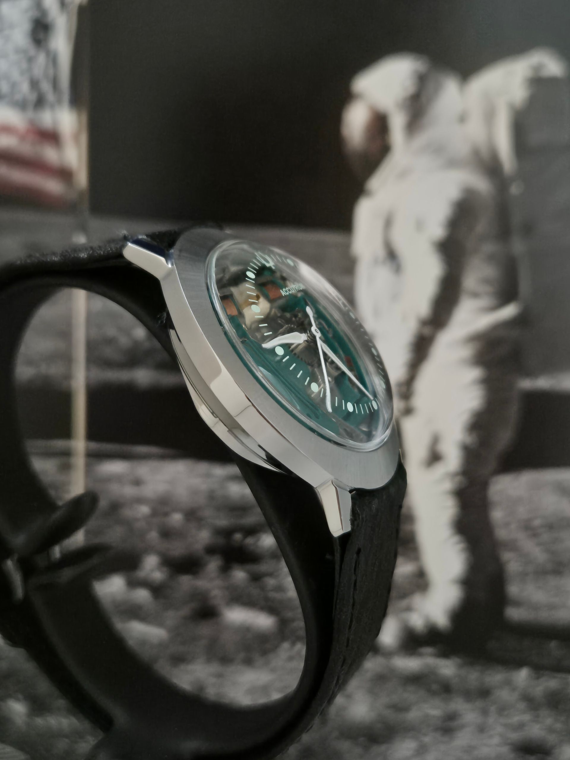 Close-up of a black wristwatch with a teal dial, worn on a person’s wrist indoors.