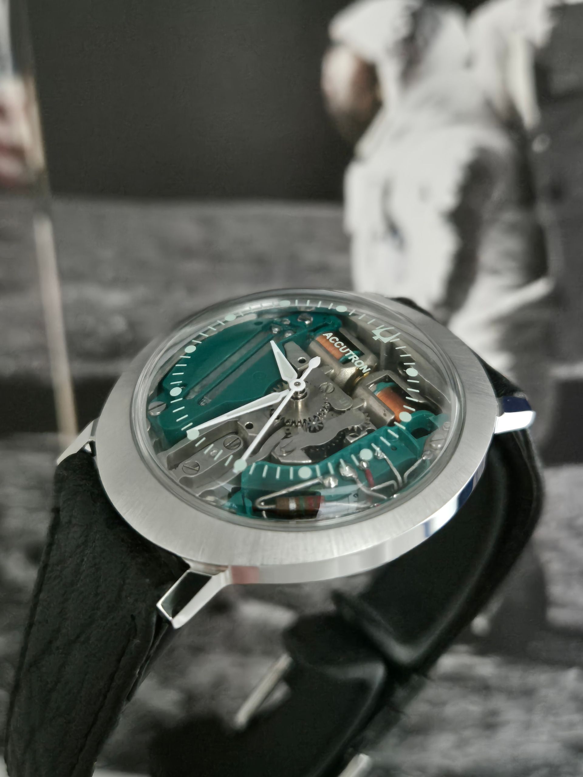 Silver wristwatch with a teal dial and black strap in close-up against a blurred black-and-white background