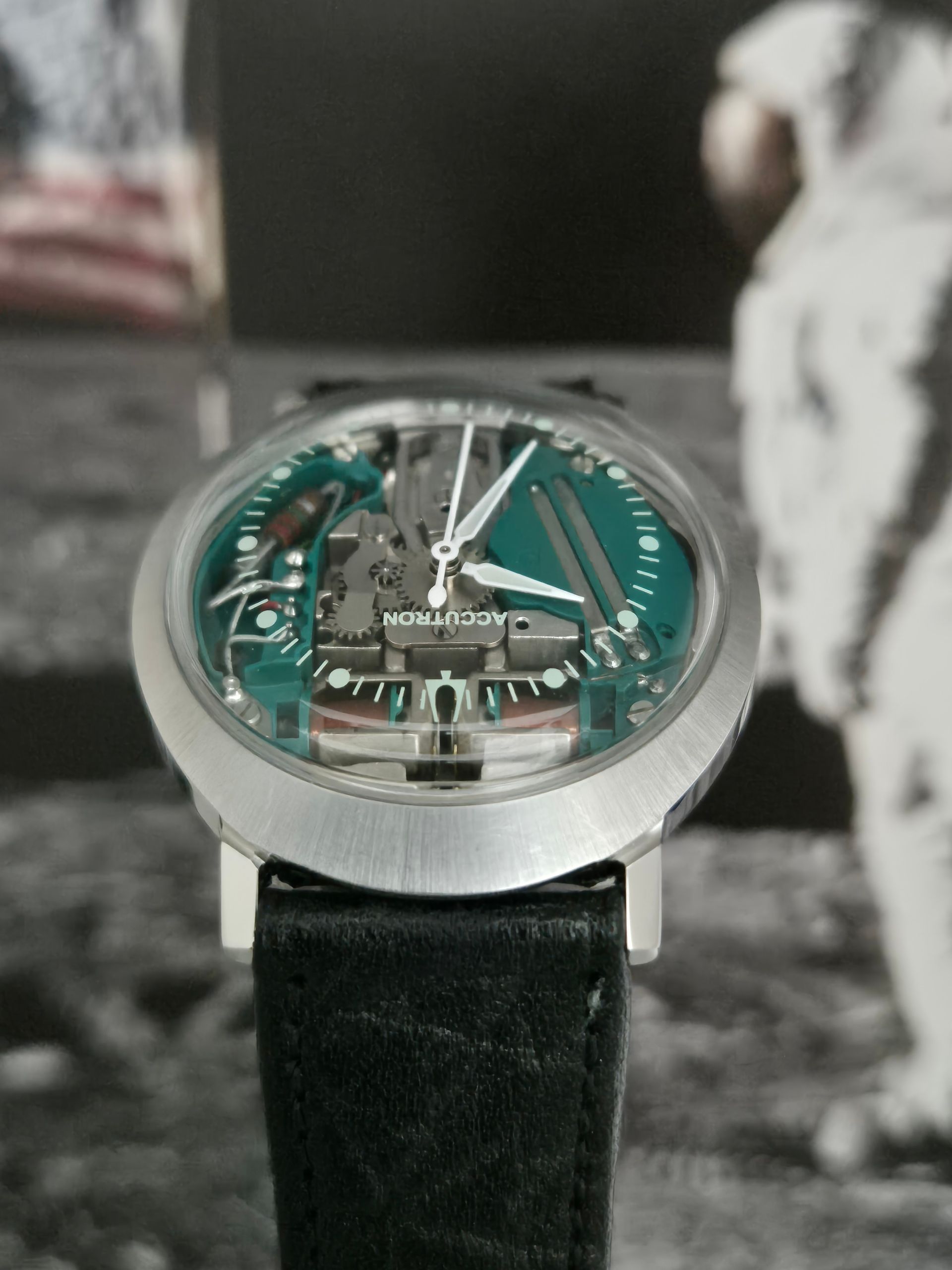 Silver wristwatch with a green skeleton dial and black leather strap on a blurred black-and-white background