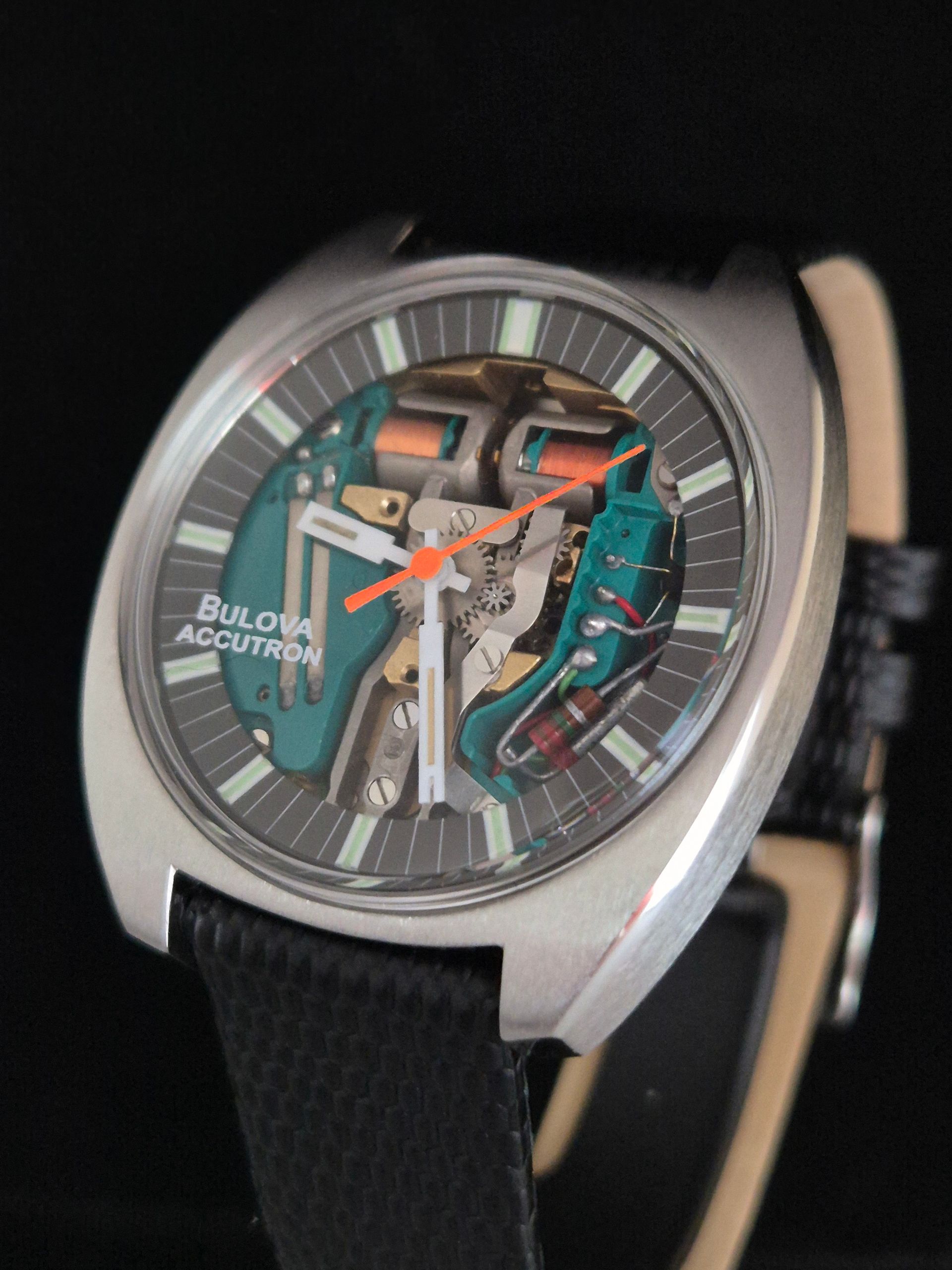 Bulova automatic wristwatch with turquoise skeleton dial, orange hands, and black strap on a dark background