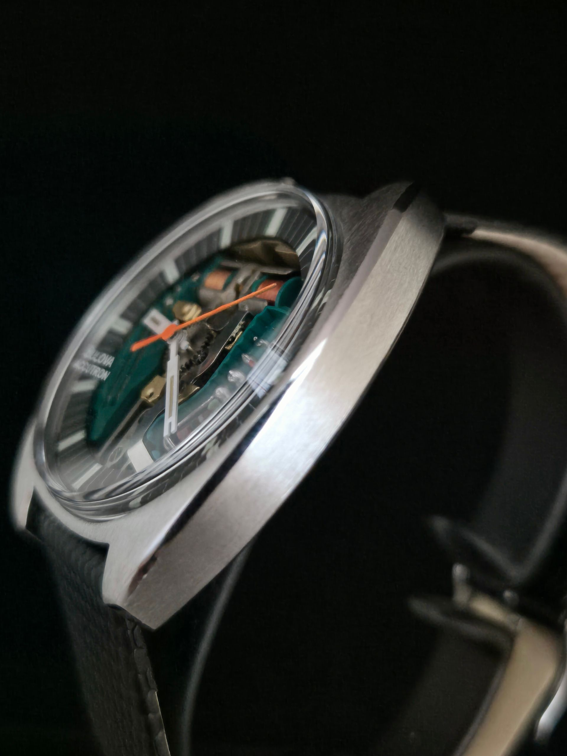 Silver wristwatch with green dial, orange hands, and black leather strap on a dark background