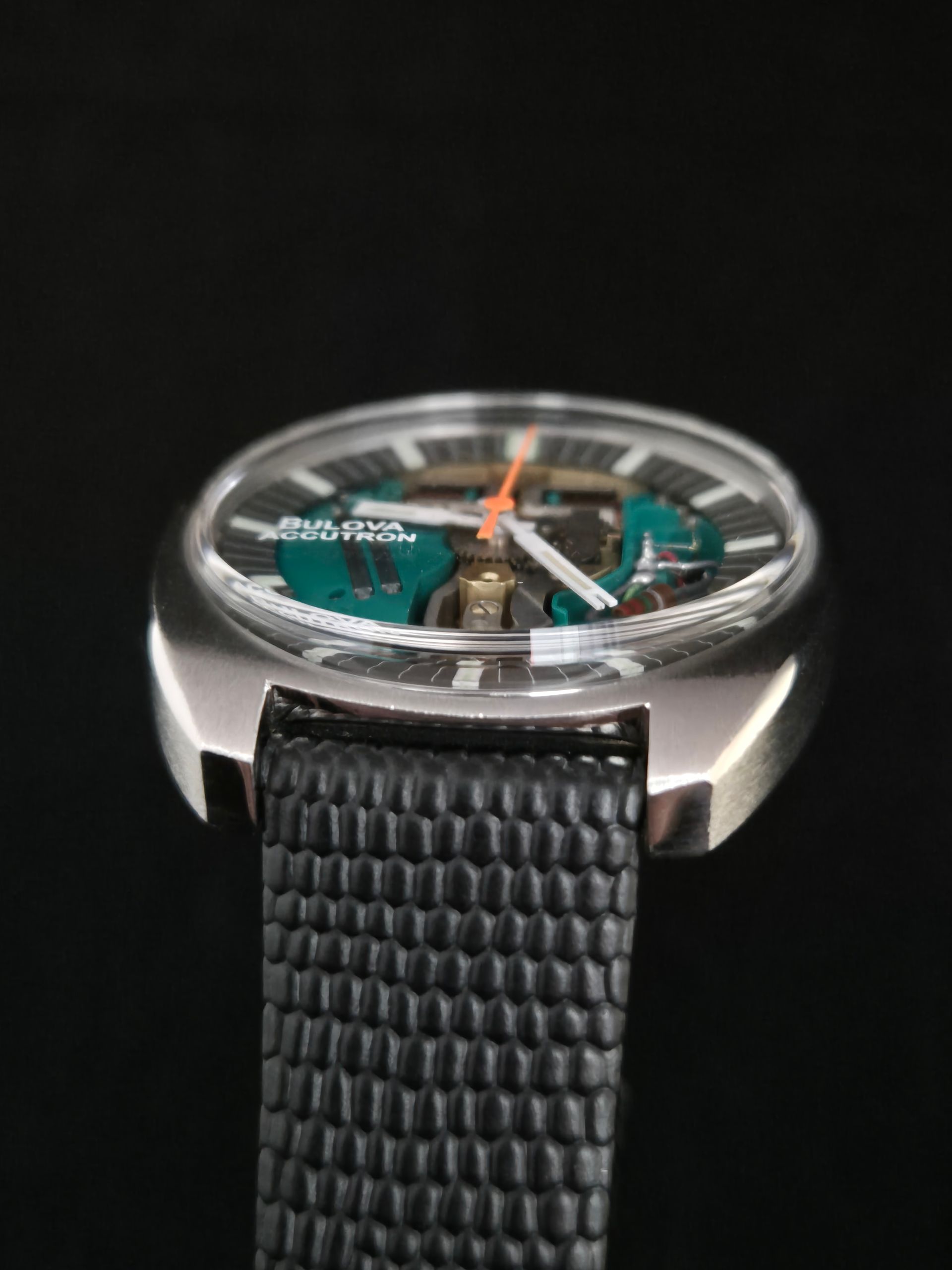 Close-up of a silver wristwatch with a black textured strap and teal dial accents on a black background