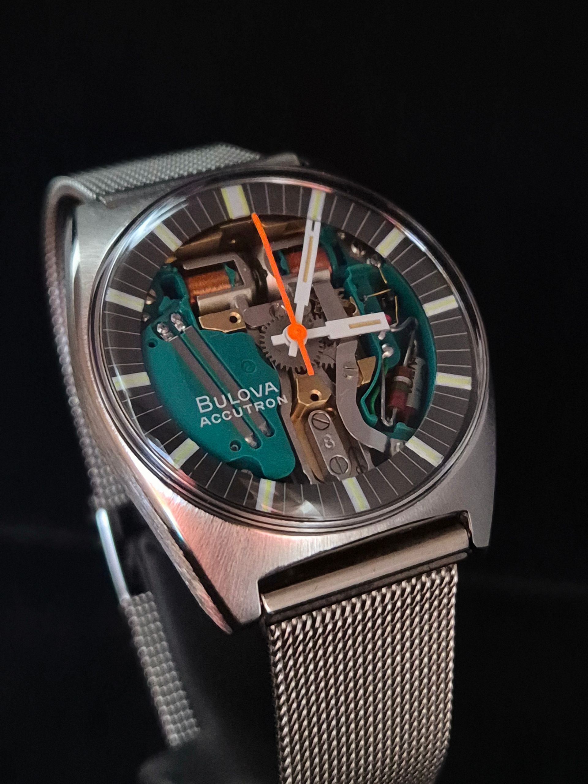 Close-up of a silver Bulova watch with turquoise open dial and orange hands on a mesh band