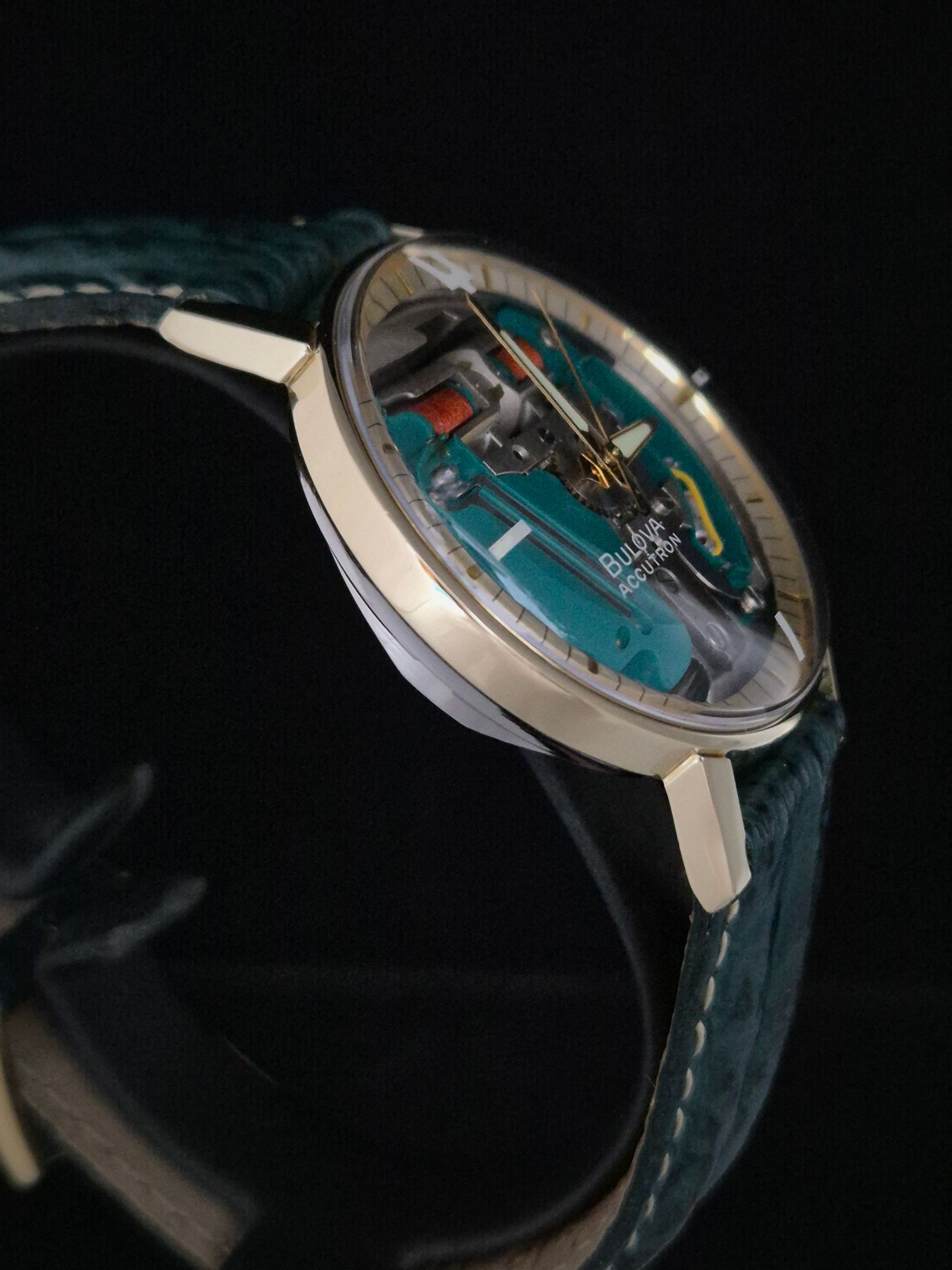 Close-up of a round wristwatch with a turquoise skeleton dial and dark leather strap on a black background