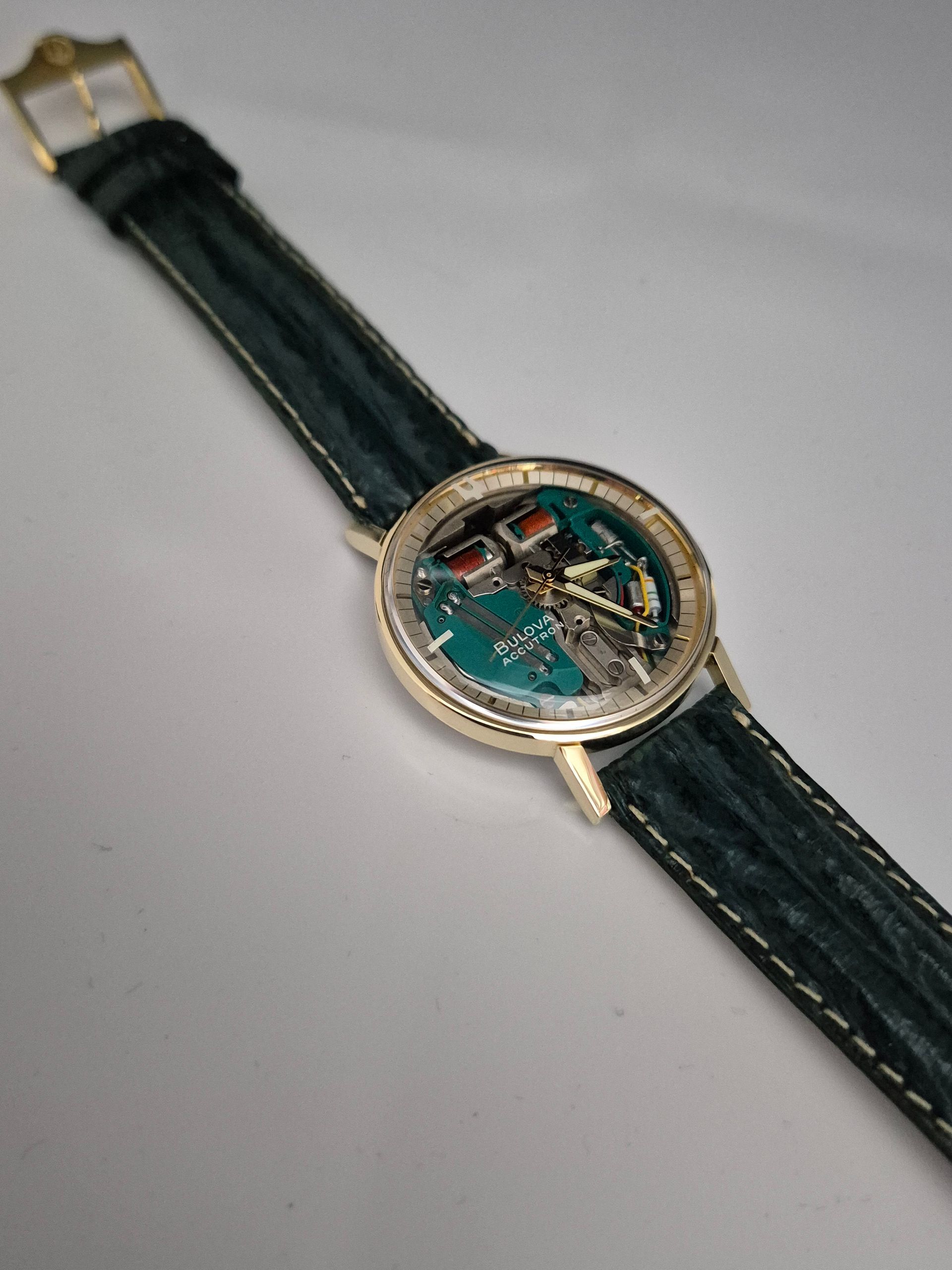 Gold wristwatch with a teal-and-black strap on a light gray surface